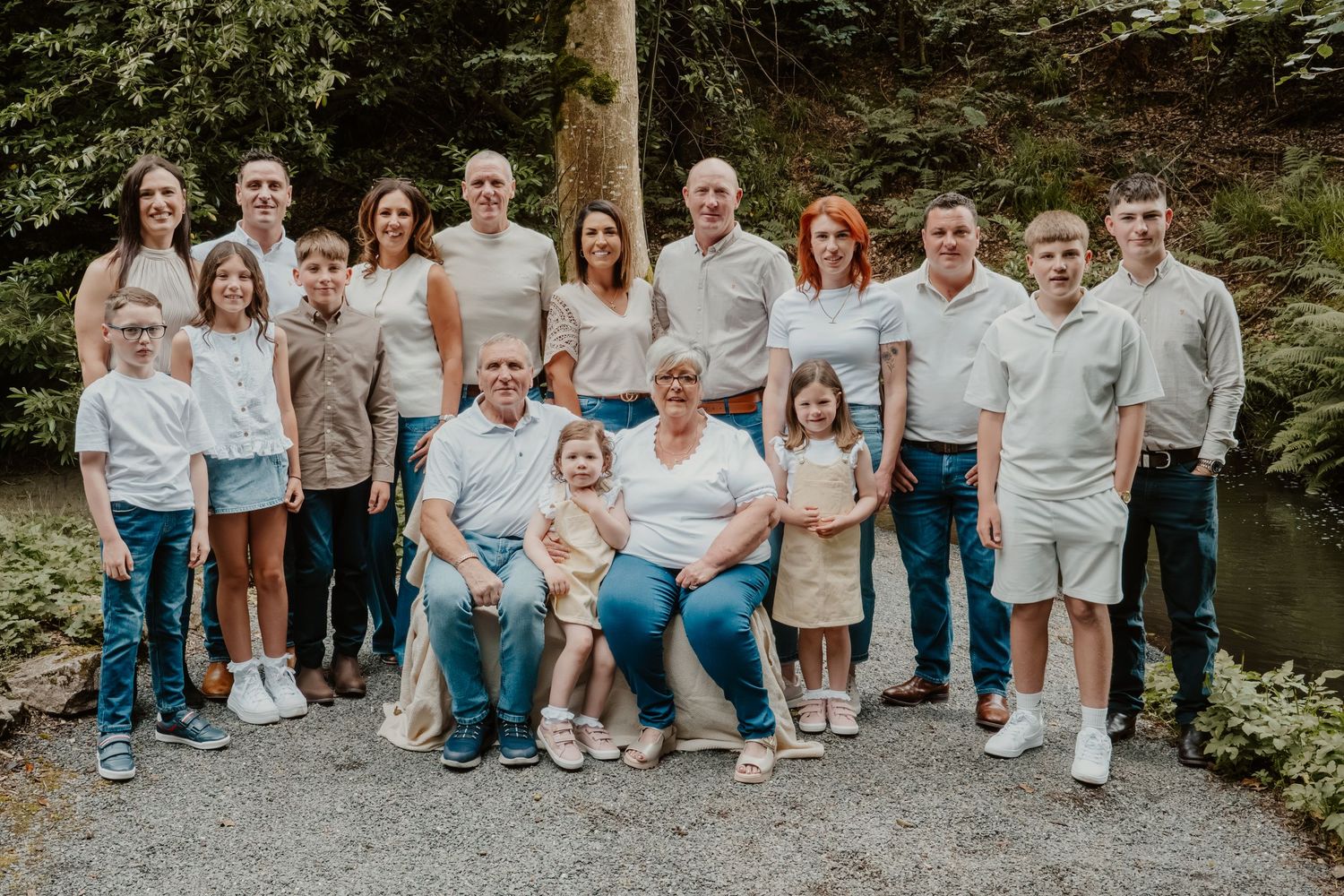 Large multigenerational family posing together outdoors near lush green trees for a formal group portrait photo.