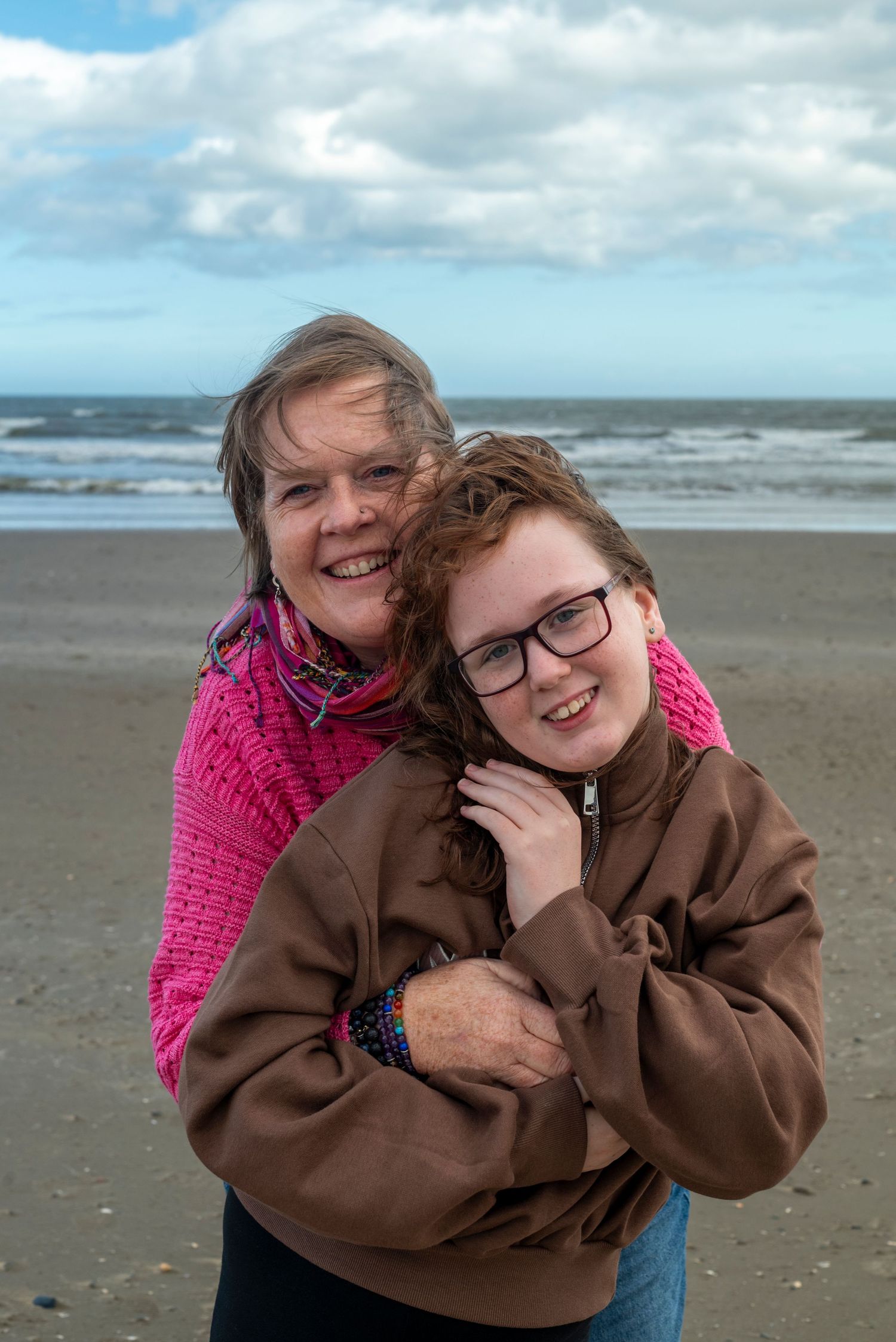Woman in pink wrap hugs smiling red-haired girl in brown hoodie on a cloudy beach.