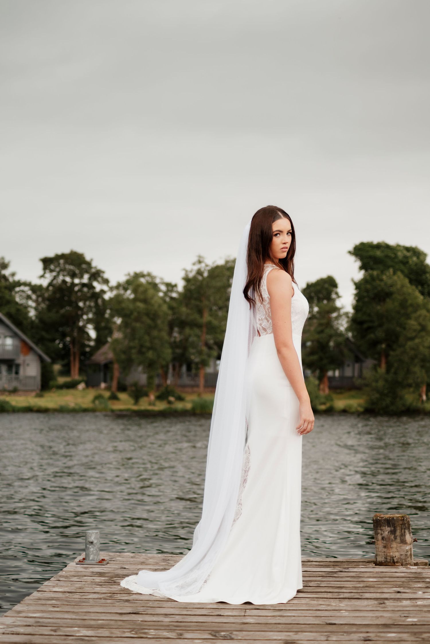 Bride in white gown and veil stands on wooden dock by lake with trees in background.