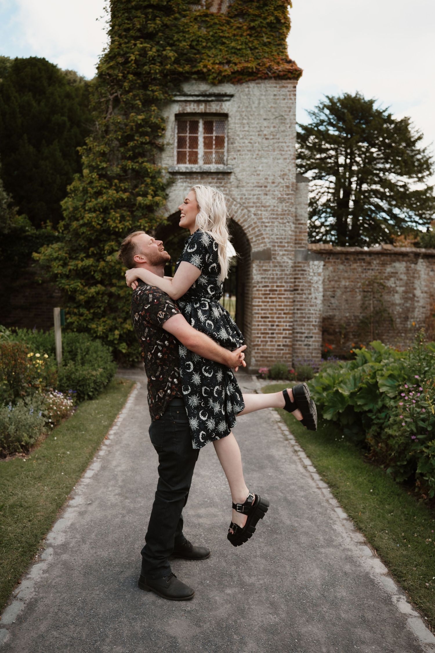 A joyful couple embracing in a romantic garden, man lifting woman in matching dark floral outfits near a historic brick tower.