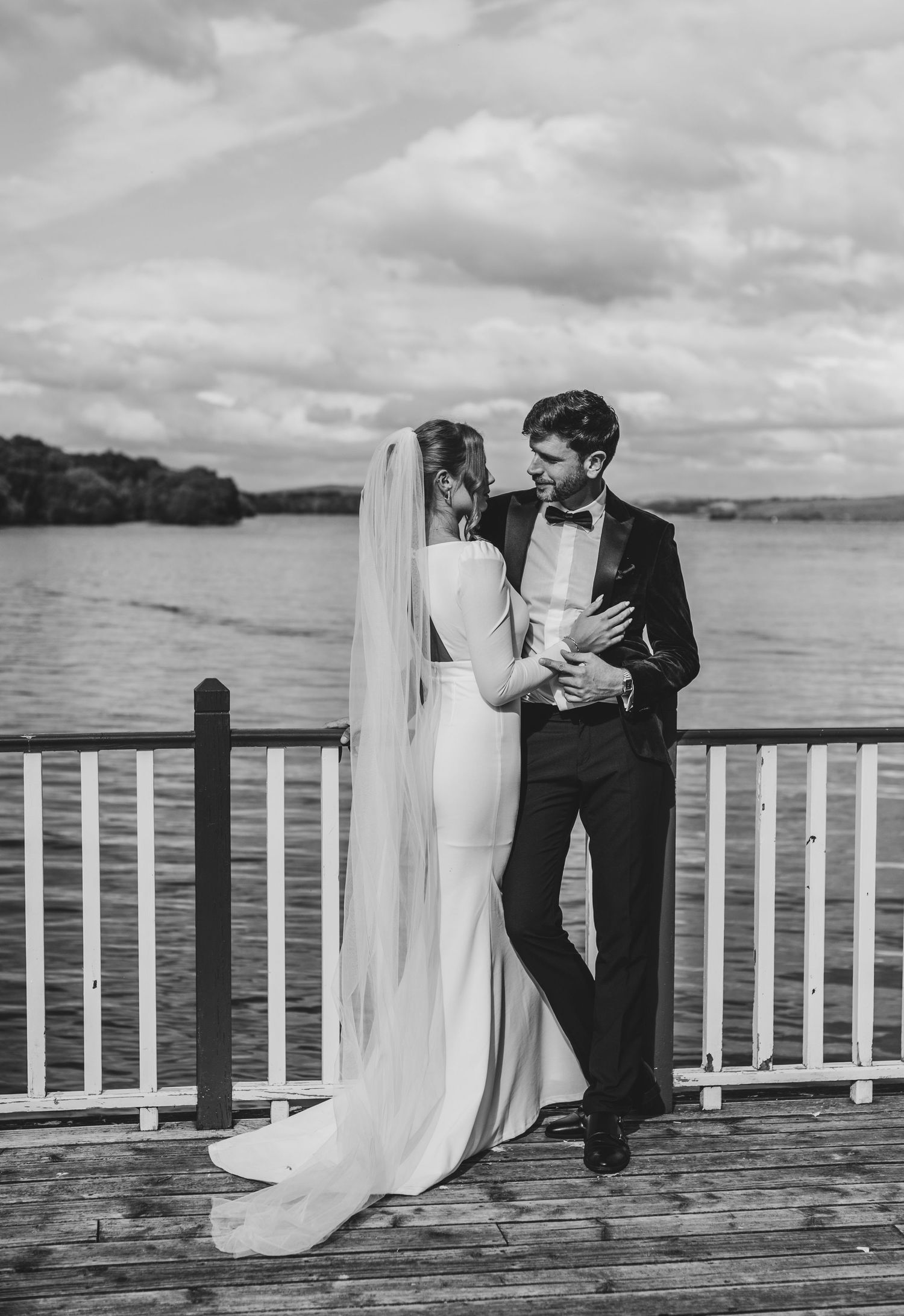 Black and white photo of bride and groom embracing on waterfront dock, bride wearing flowing veil.