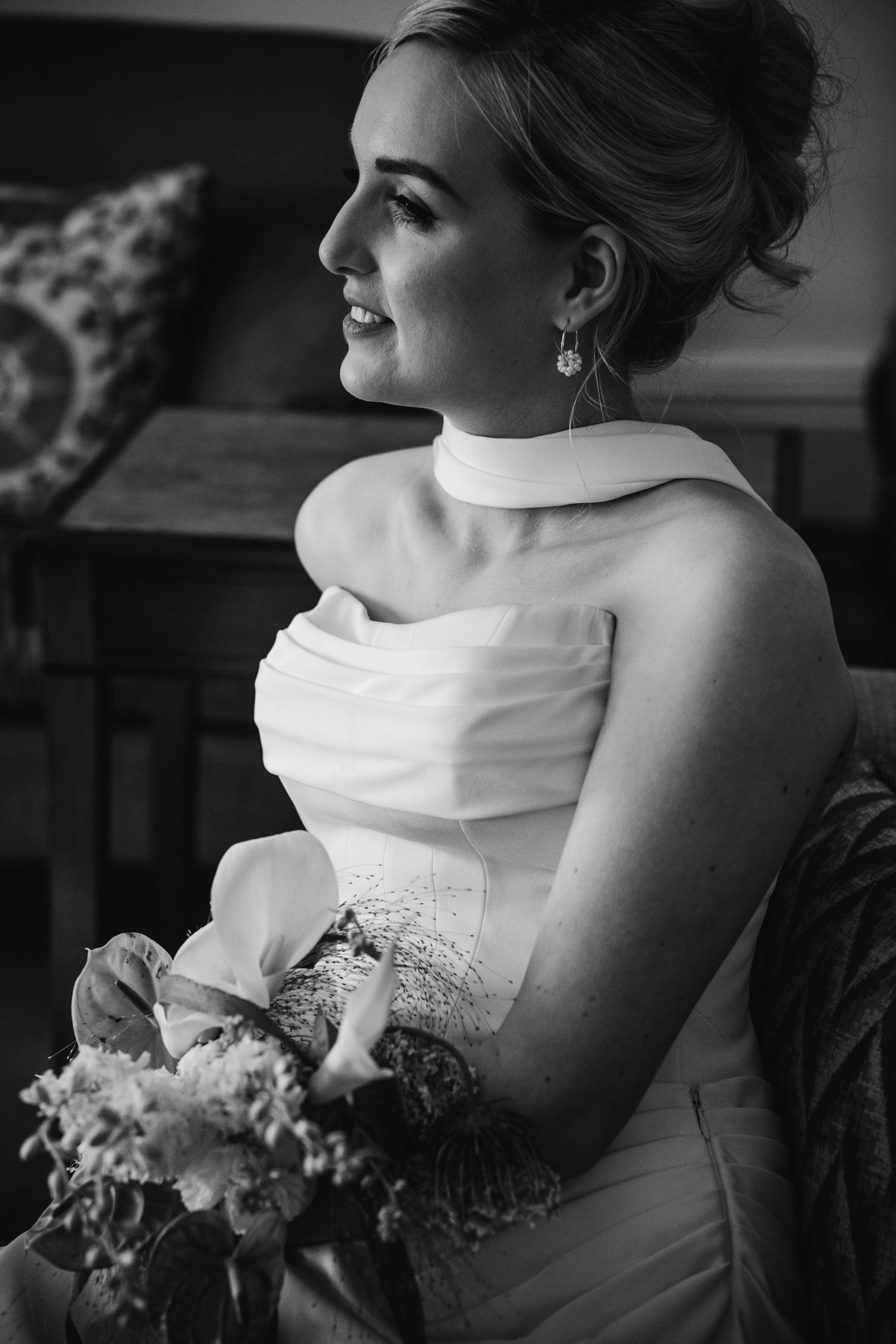 Smiling bride in strapless draped white gown holding floral bouquet seated indoors in black and white photo.