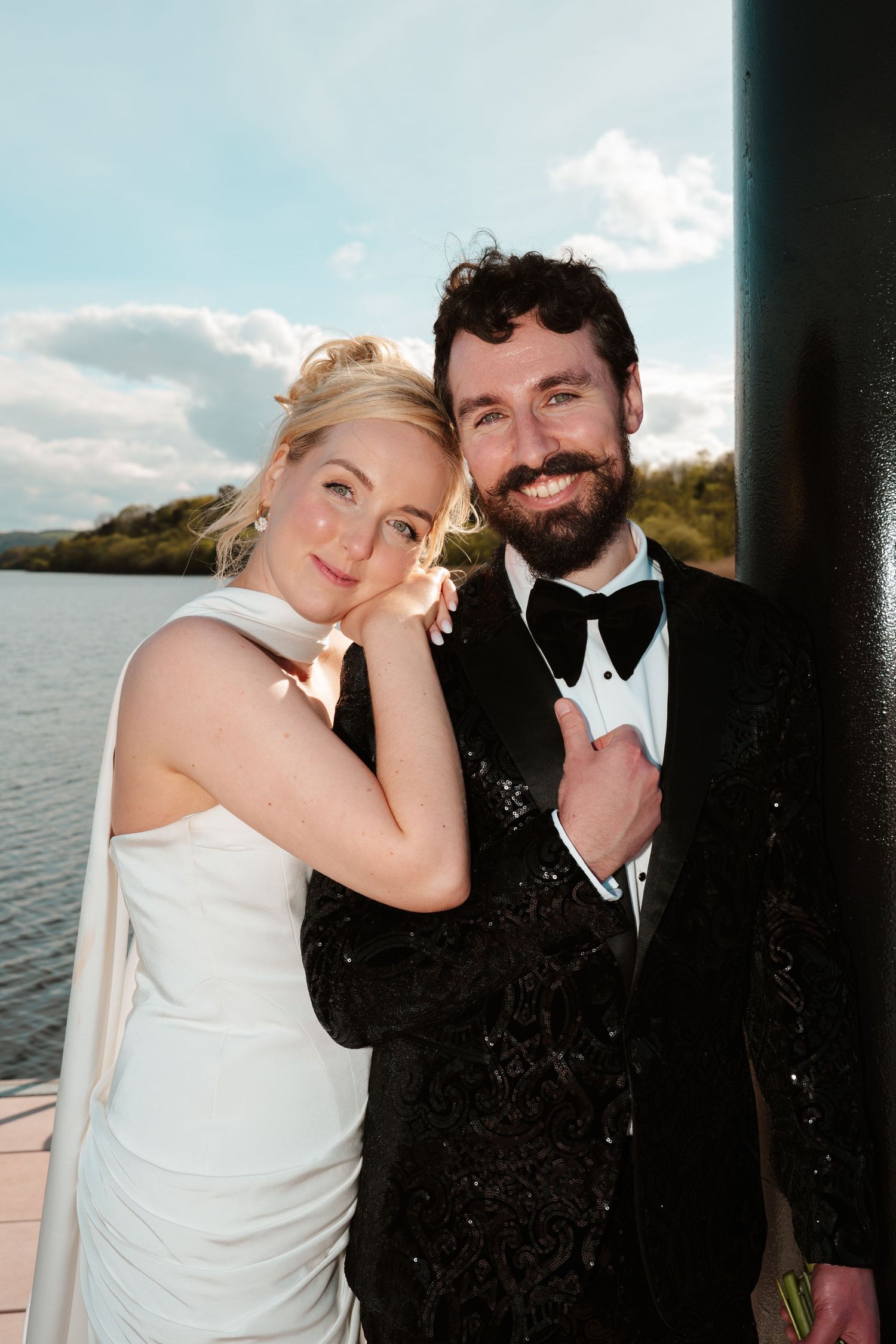 Smiling bride in white gown leans on bearded groom in black tuxedo by waterfront.
