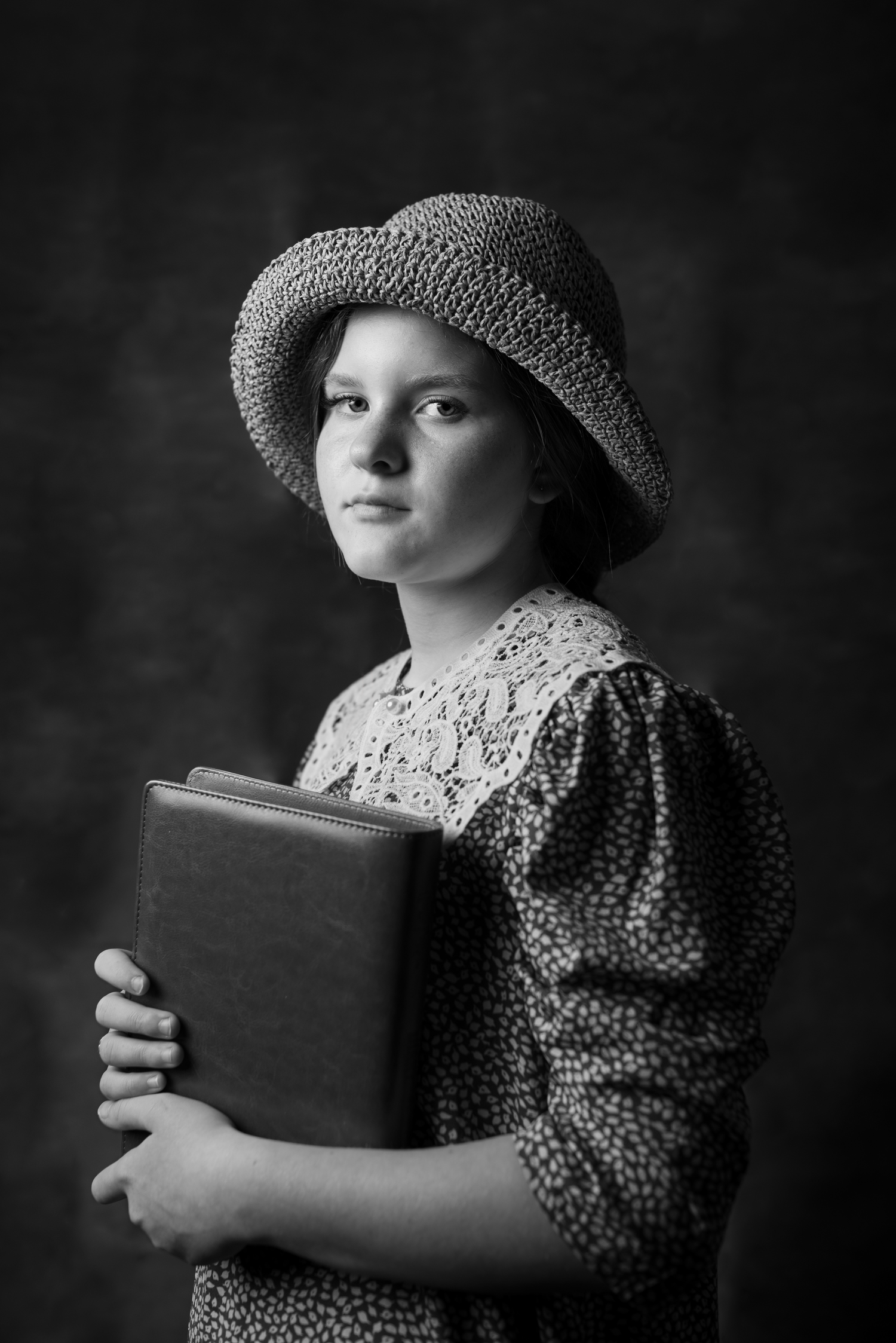Black and white portrait of a young girl in a floral dress, lace collar, and straw hat holding a book.
