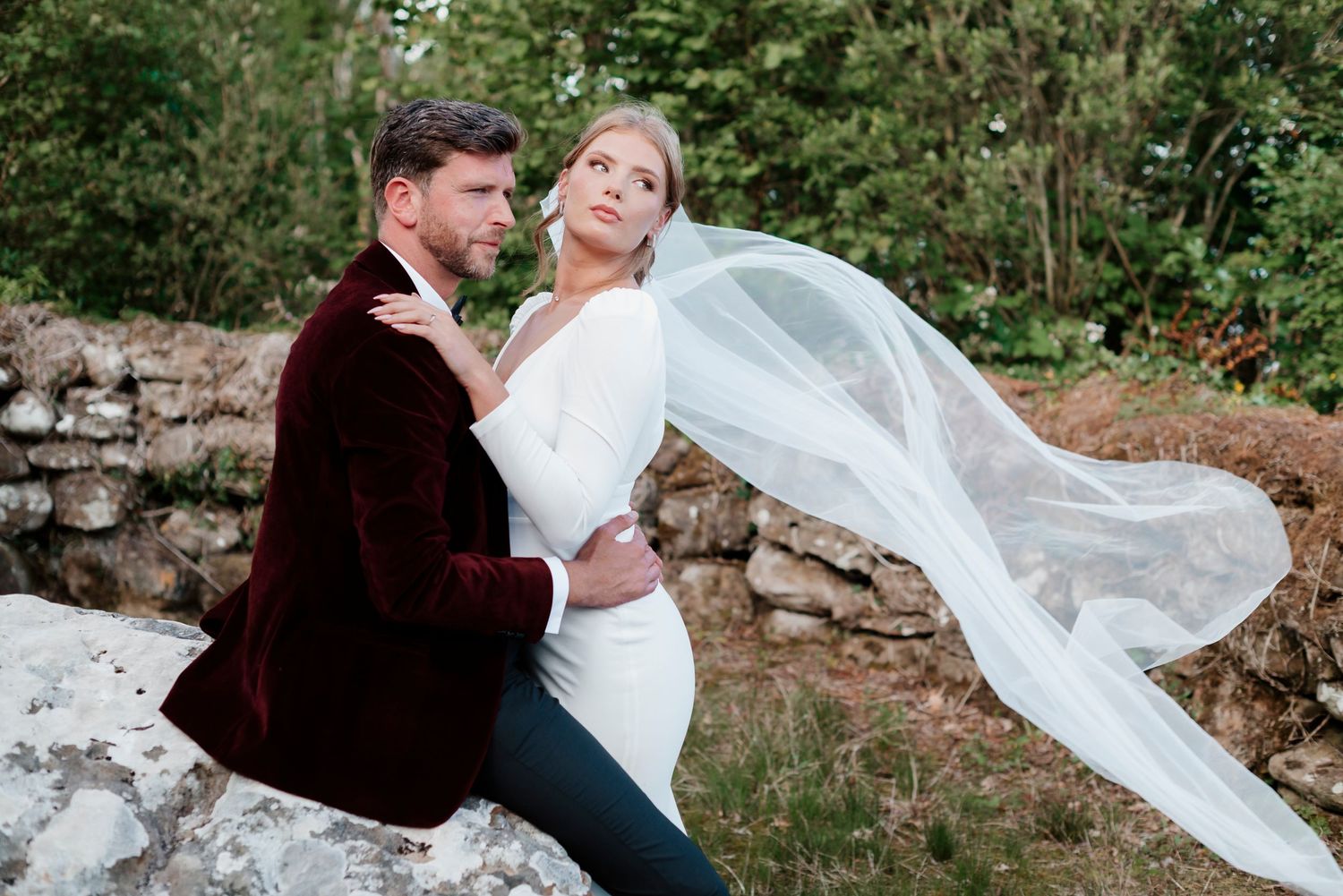 Groom in burgundy velvet jacket holds bride as her veil flows in wind by rustic stone wall.