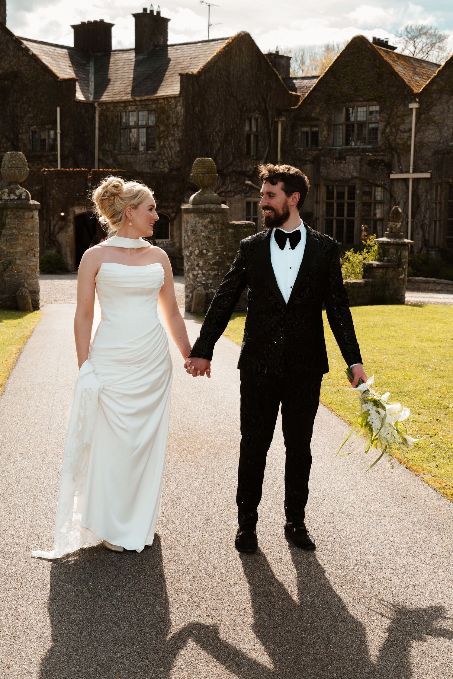 Bride and groom hold hands walking past a historic stone manor house on their wedding day.