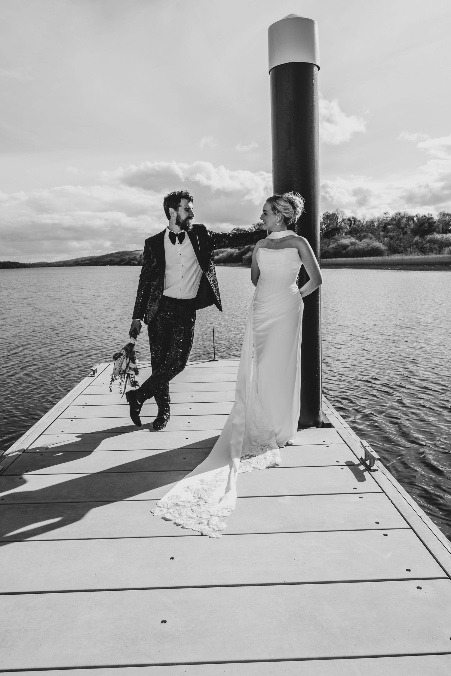 Bride and groom pose on a lakeside dock in a stunning black and white wedding portrait.