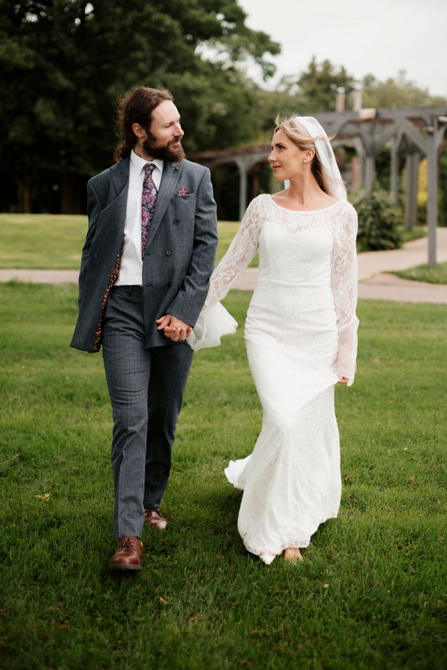 Bride and groom hold hands walking on green lawn in wedding attire outdoors.