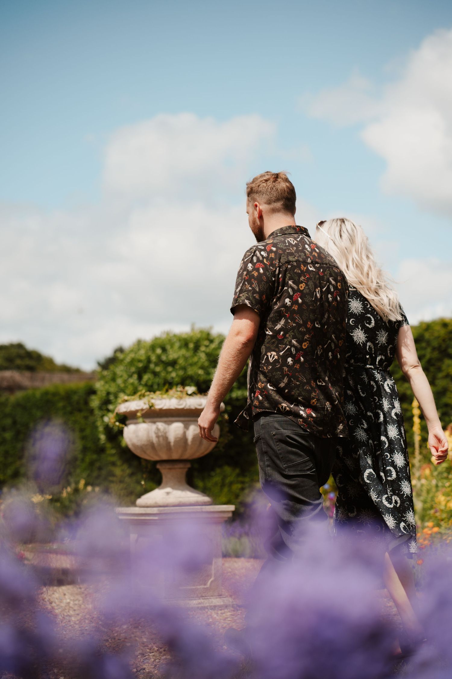 A couple holding hands and walking through a beautiful garden with purple lavender blooms and a stone urn planter.