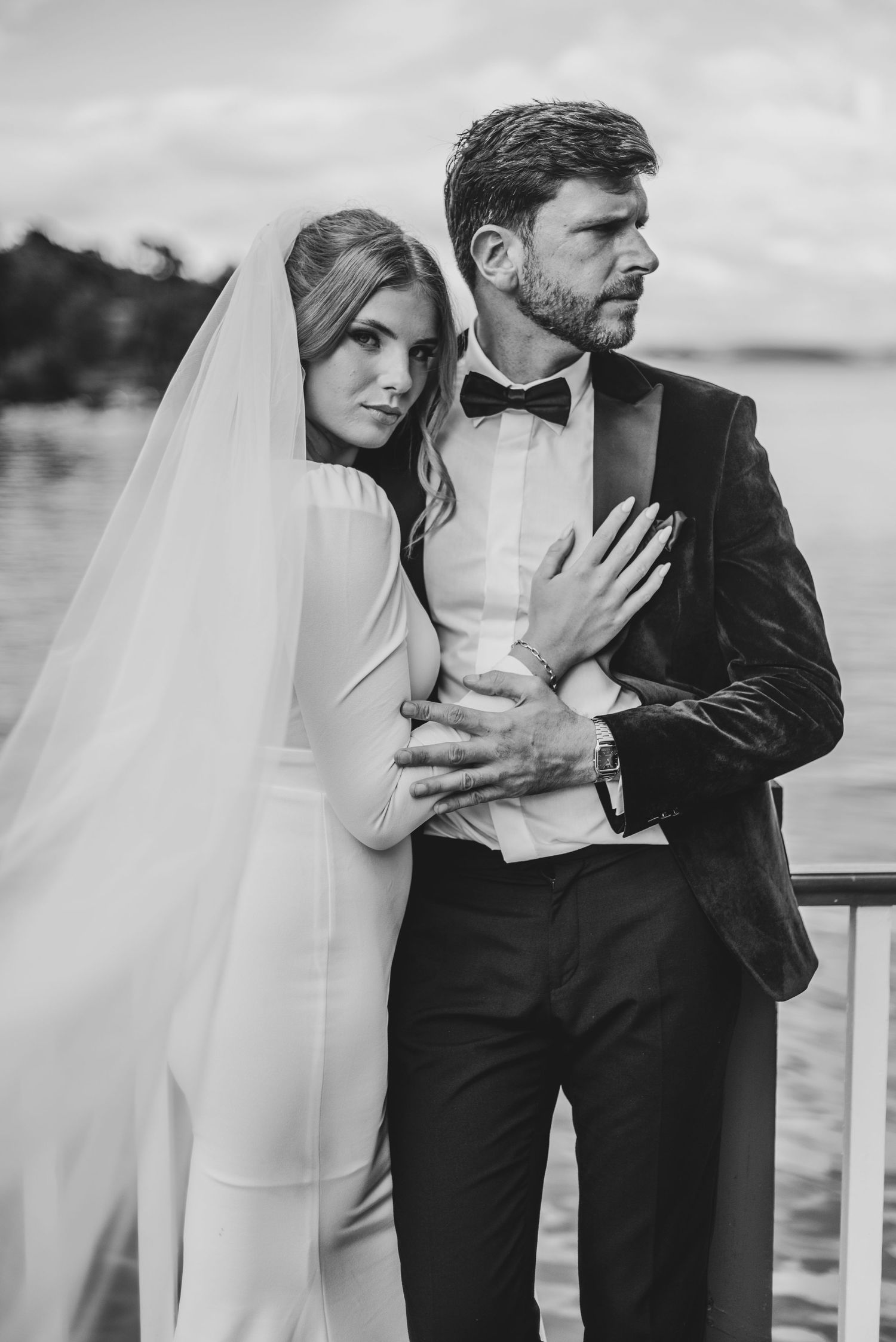 Elegant bride in veil embraces groom in velvet tuxedo on waterfront balcony.