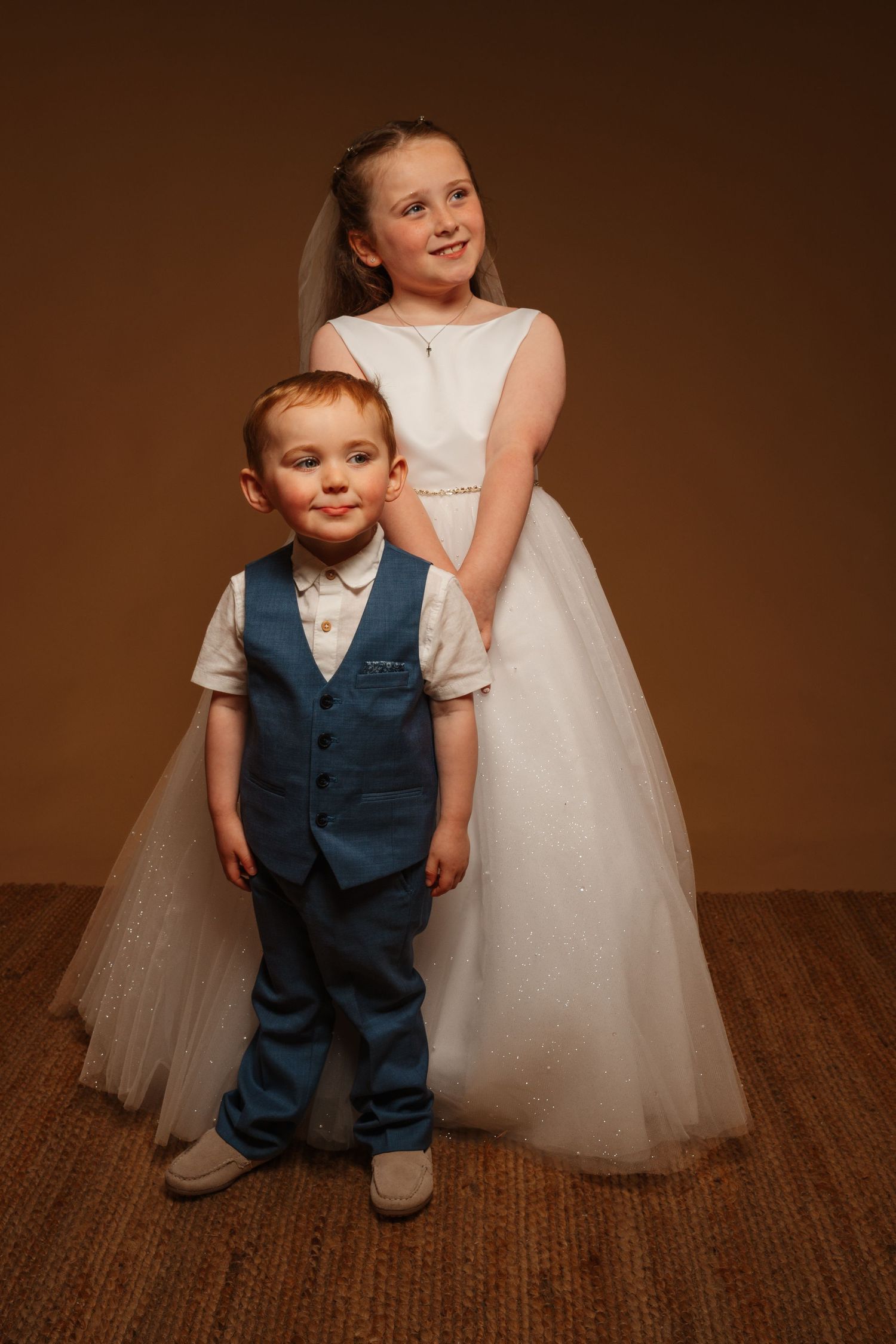 Young girl in white communion dress with veil stands behind smiling boy in blue suit for formal portrait.