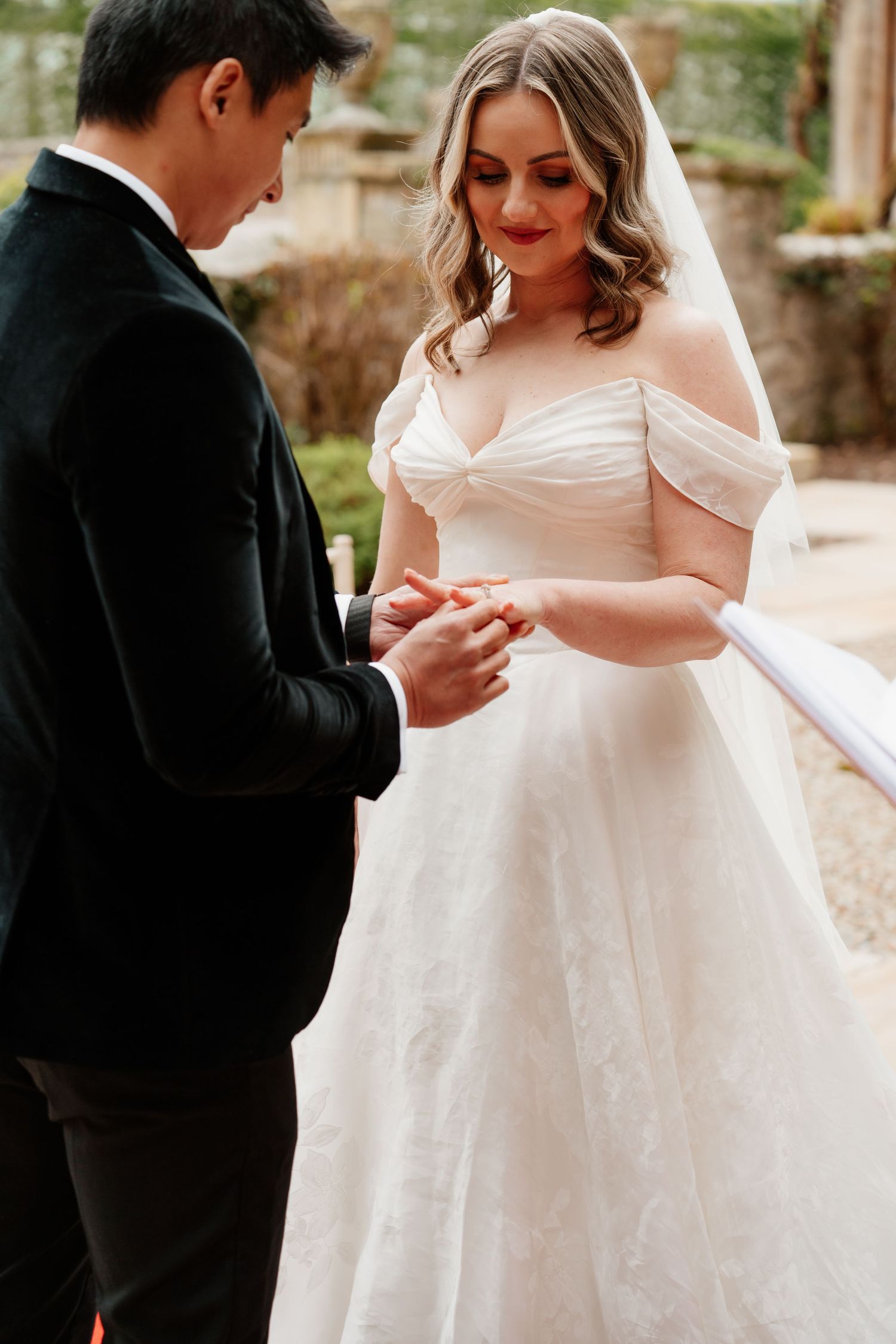Groom placing wedding ring on brides finger during outdoor ceremony, bride in off-shoulder ball gown.