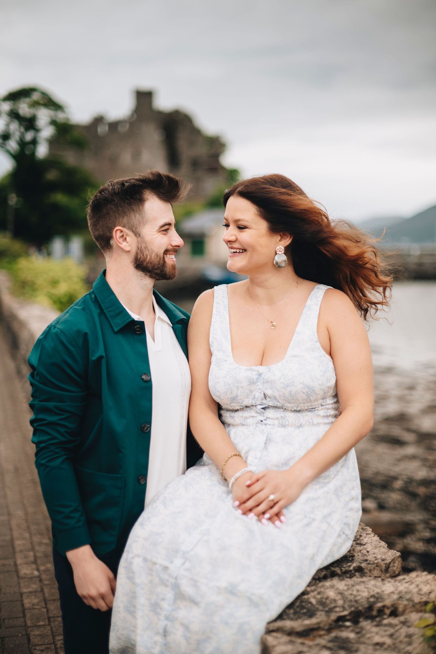 Happy couple sharing a romantic moment seated on a stone wall near the water with a historic castle behind them.