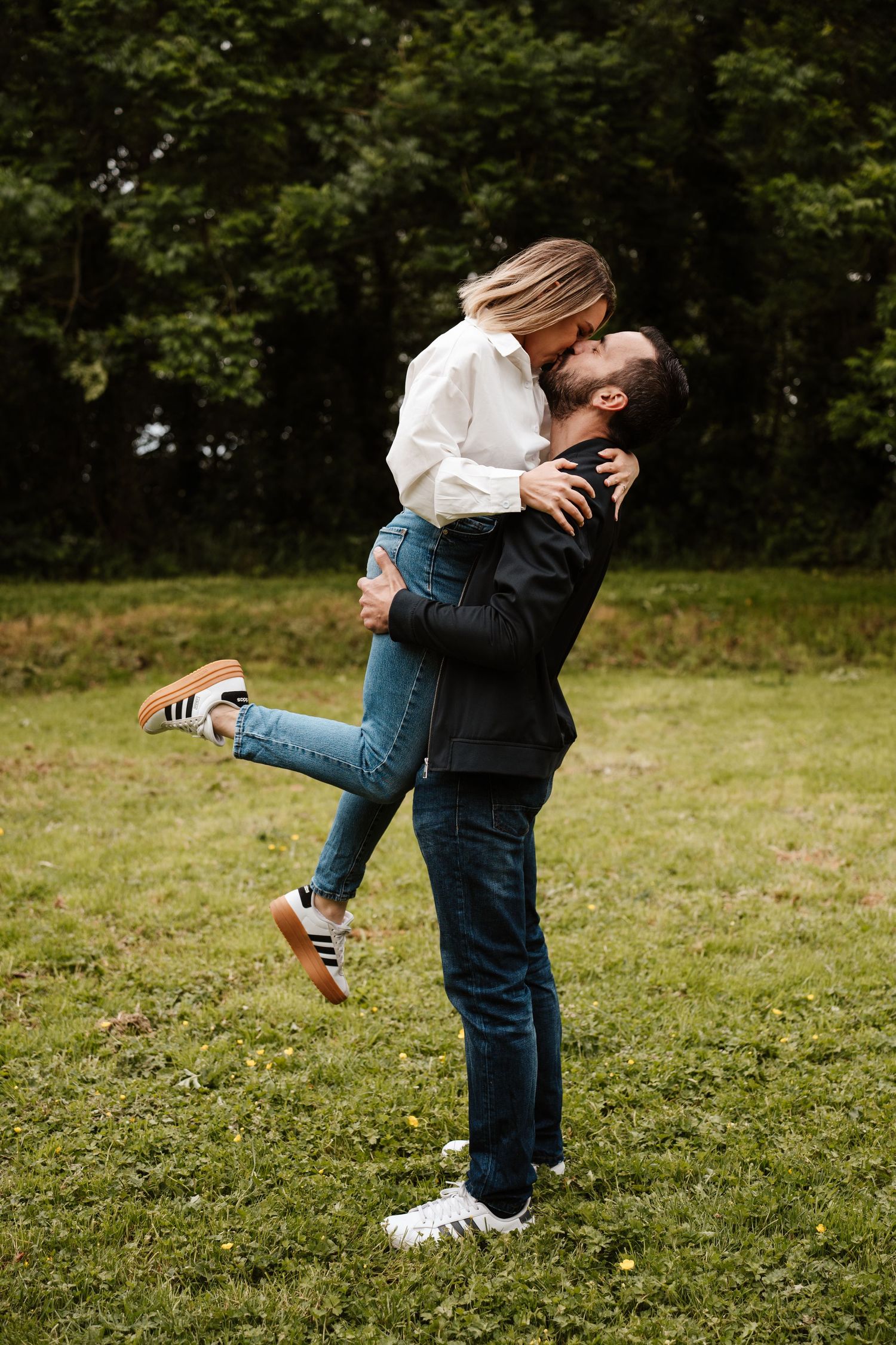 Man lifting woman for a kiss in a lush green park, both wearing jeans and casual tops.