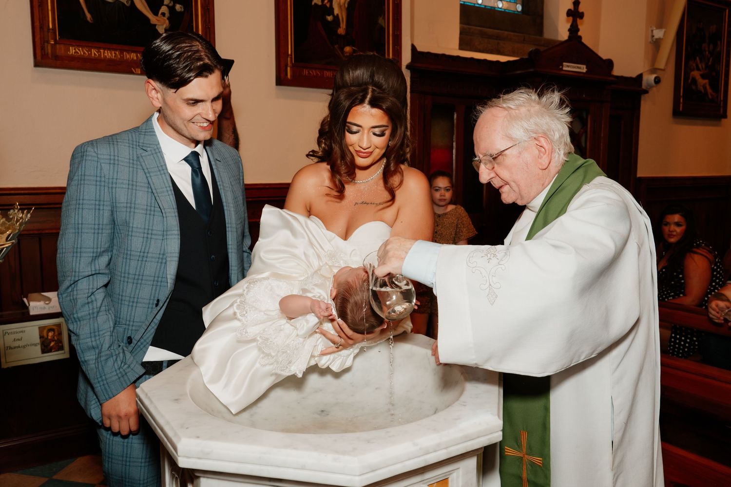 Priest performing a Catholic baptism ceremony, pouring holy water over infant at stone font with parents standing beside.