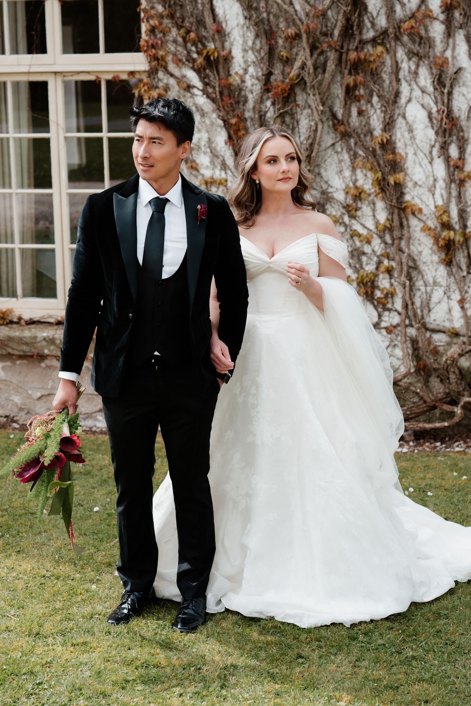 Bride in off-shoulder white gown and groom in black tuxedo holding burgundy bouquet pose outdoors at wedding.