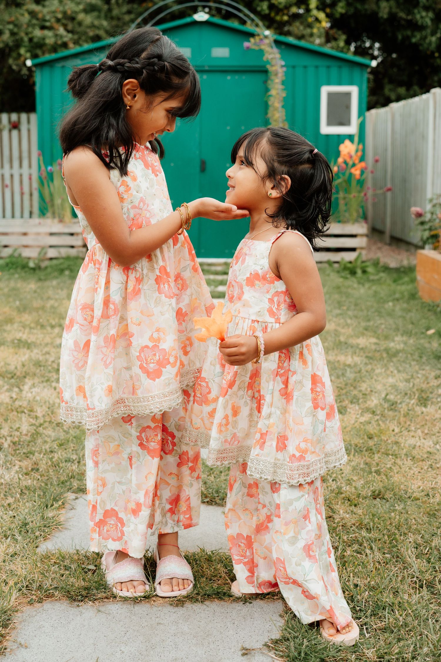 Two young sisters in matching floral outfits share a sweet moment in a sunny backyard garden.