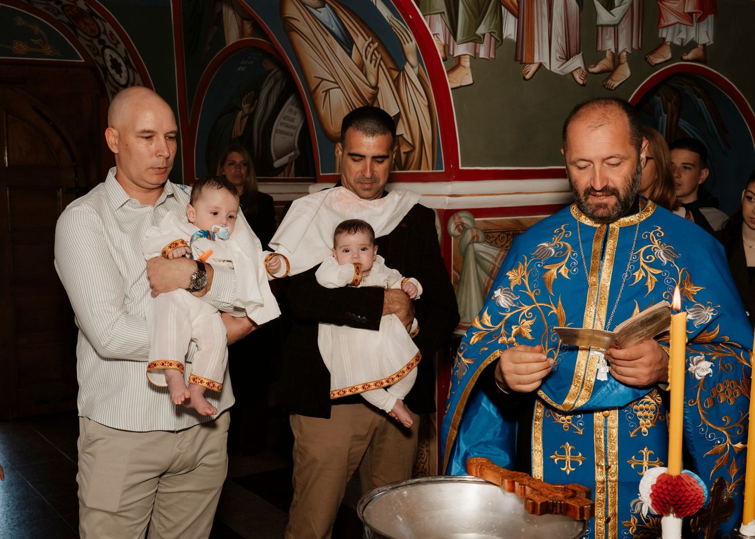Orthodox priest in blue vestments performs baptism ceremony with two men holding infants in a decorated church.