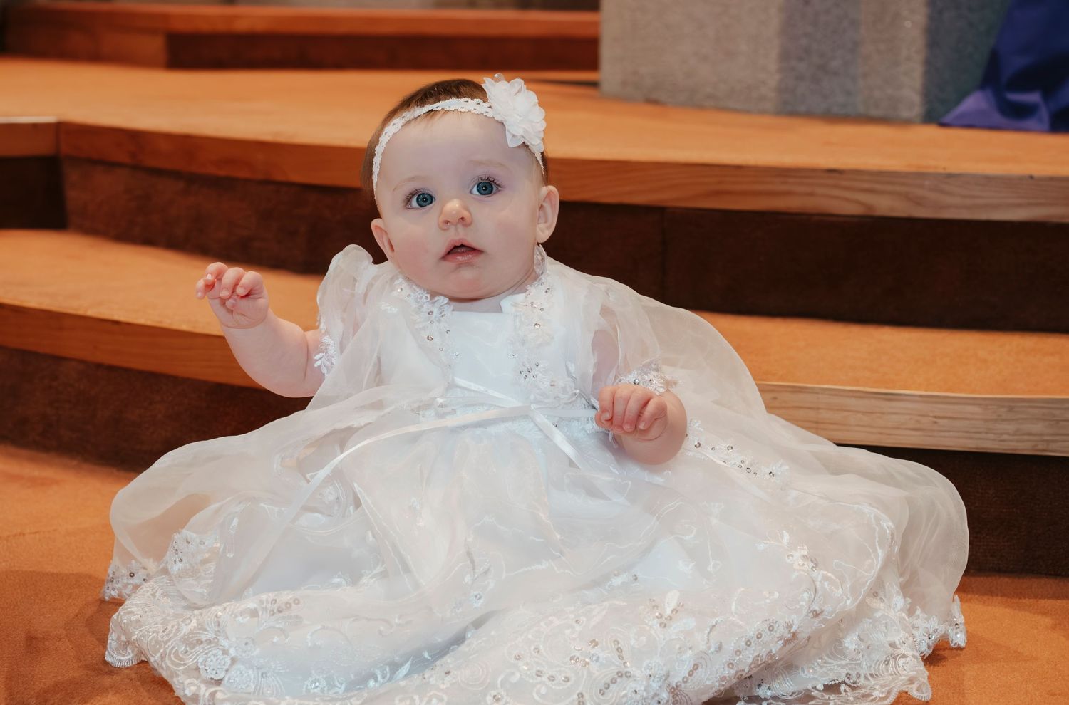 Adorable baby girl in white lace christening gown sitting on church steps with a floral headband.