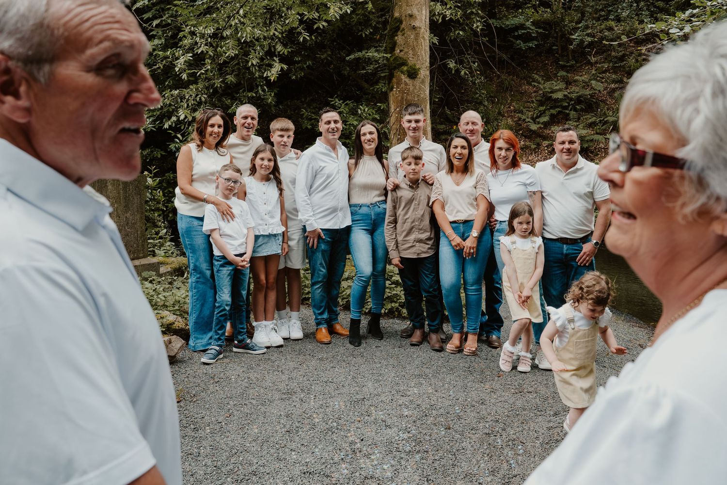Large multigenerational family group gathered outdoors on a gravel path surrounded by lush green trees.