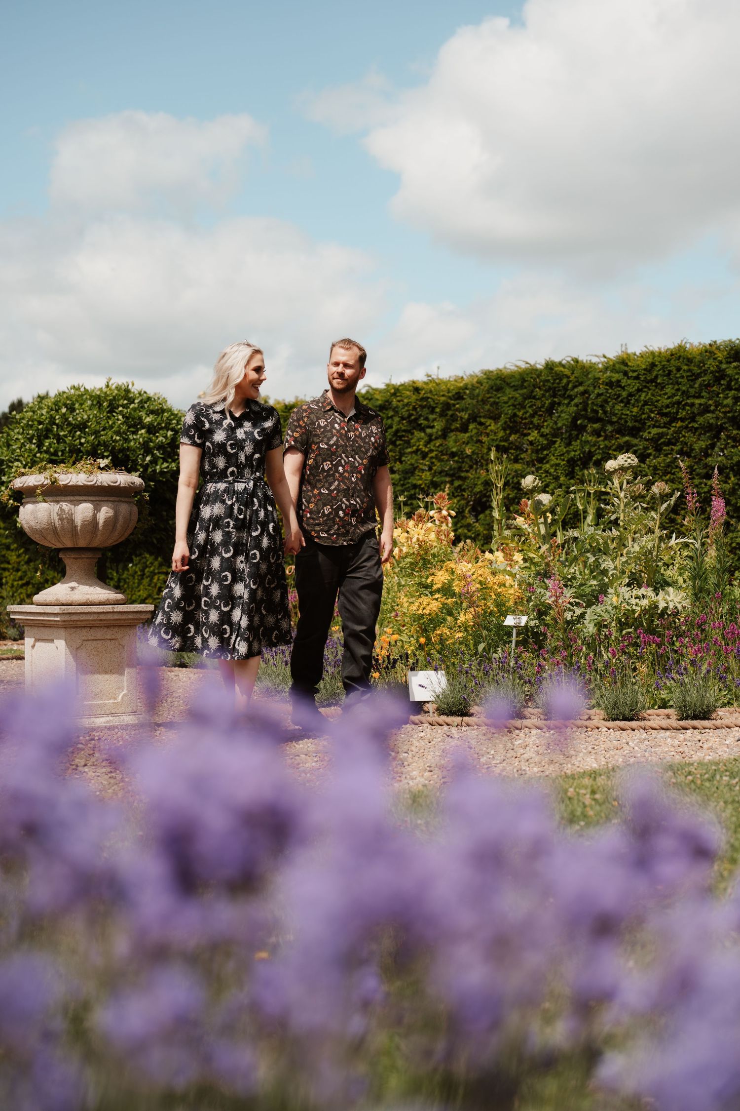 Couple walking hand in hand through a formal garden with purple lavender blooms and stone urns.