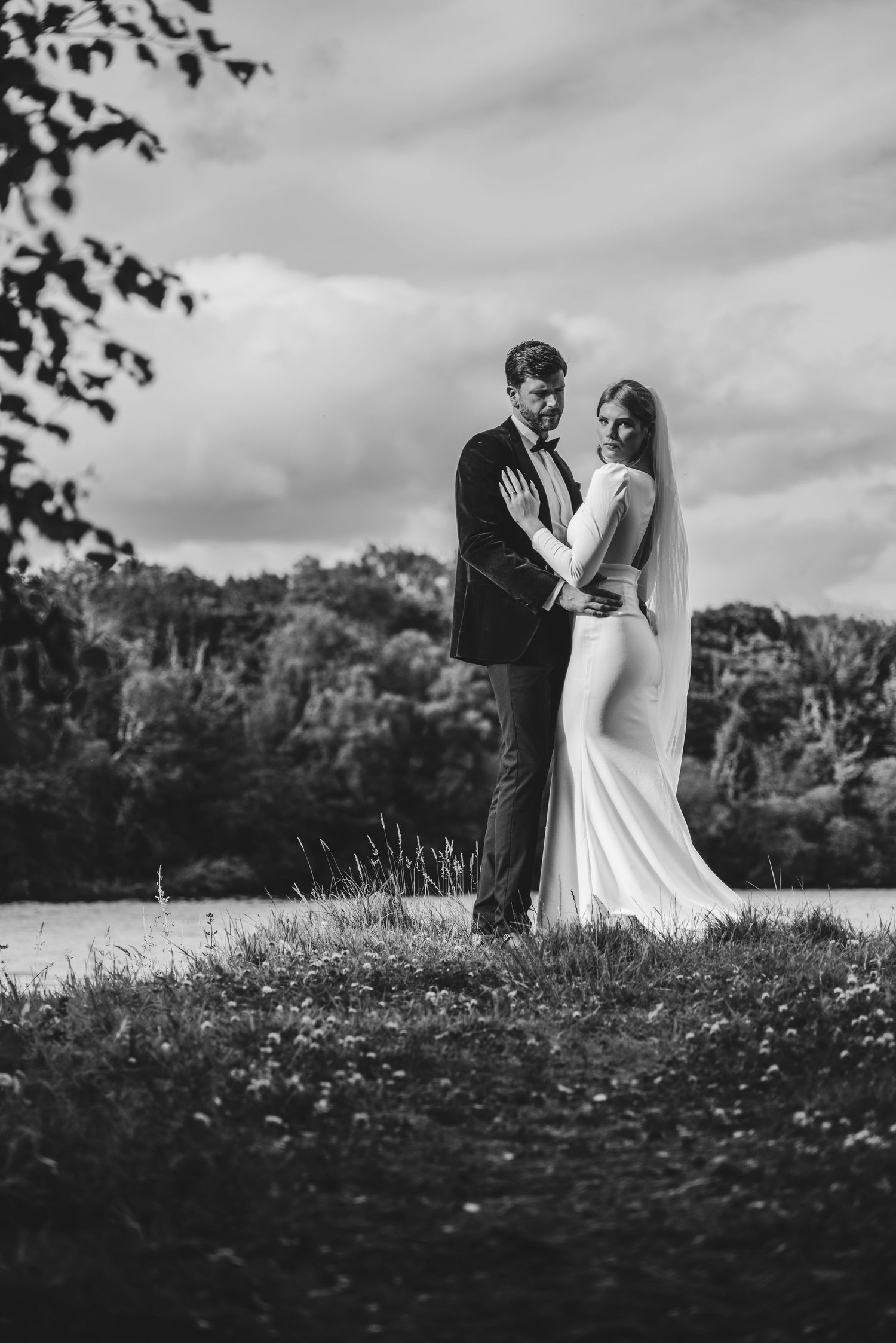 Black and white photo of bride in fitted gown with veil embracing groom outdoors near scenic lake.