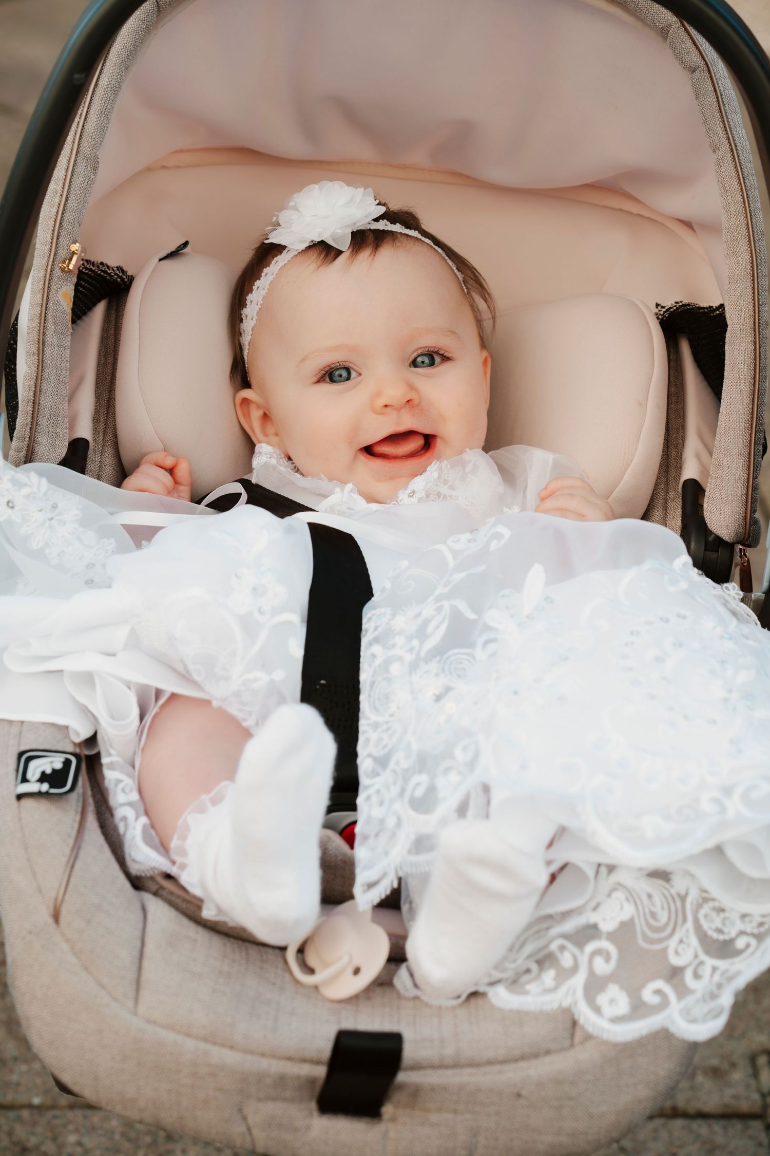 Adorable smiling baby girl in white lace christening gown seated in infant carrier.