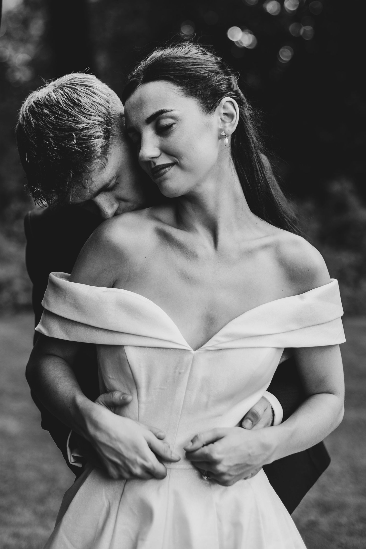 Black and white photo of a groom embracing bride in off-shoulder wedding dress, sharing a tender romantic moment outdoors.