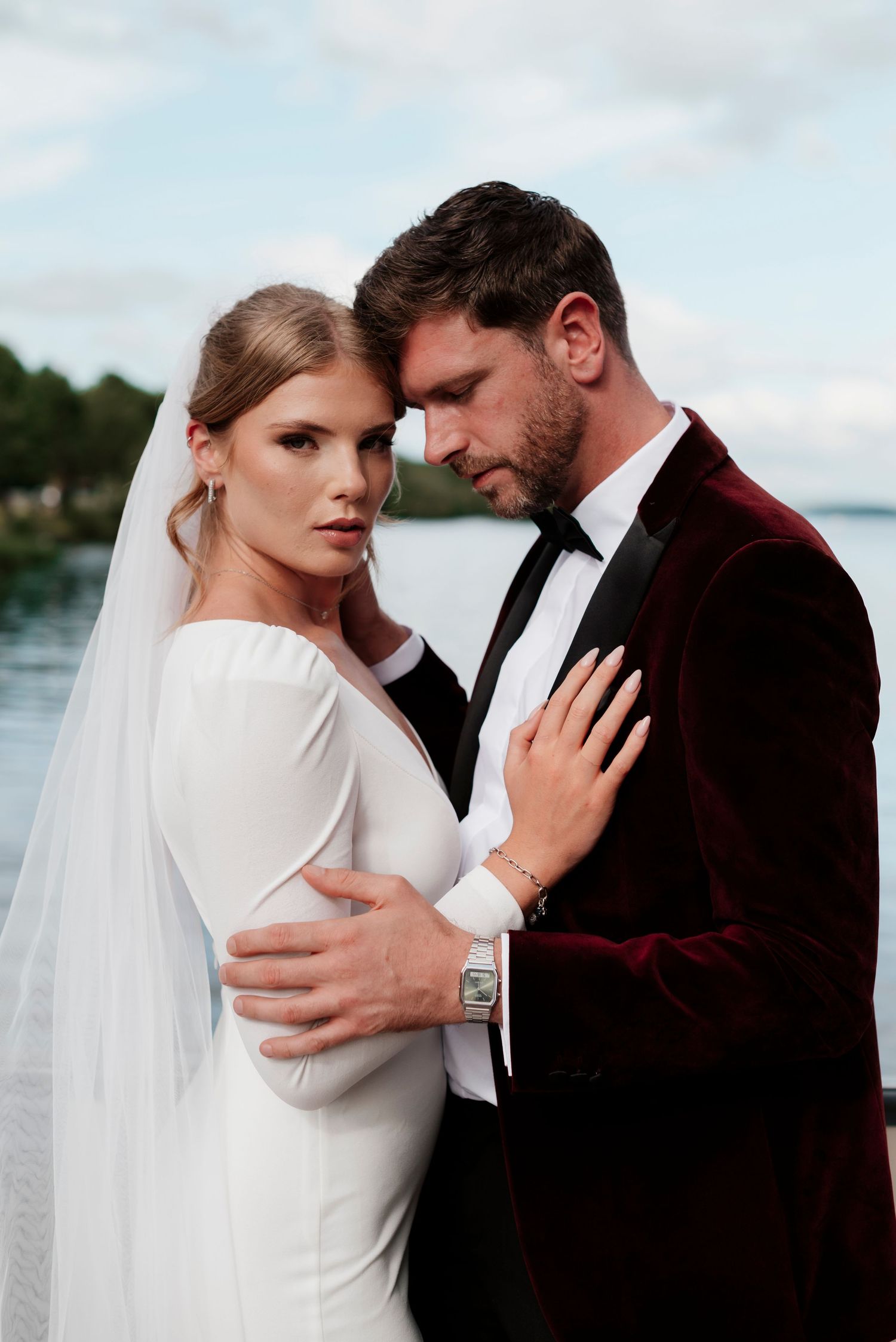 Bride in white gown and veil embraces groom in burgundy velvet tuxedo by a lakeside.