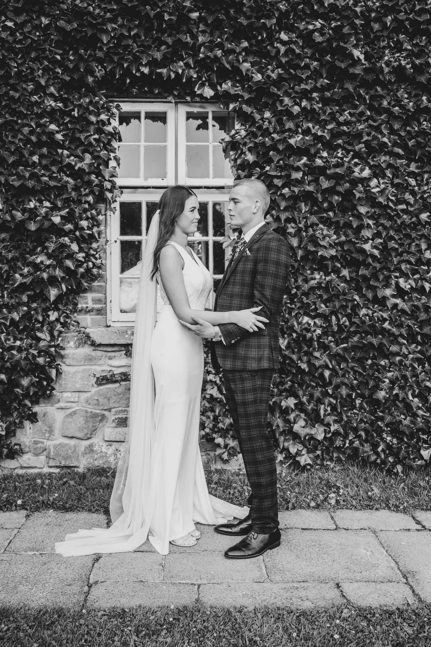 Black and white photo of bride and groom embracing before ivy-covered stone wall.