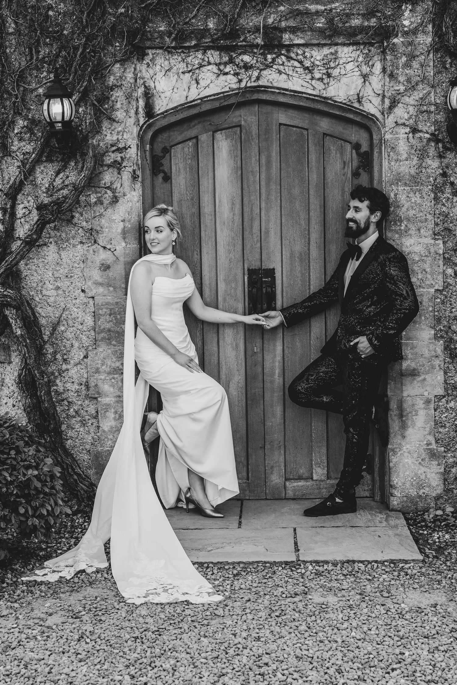 Black and white wedding photo of bride and groom holding hands by a rustic arched wooden door.