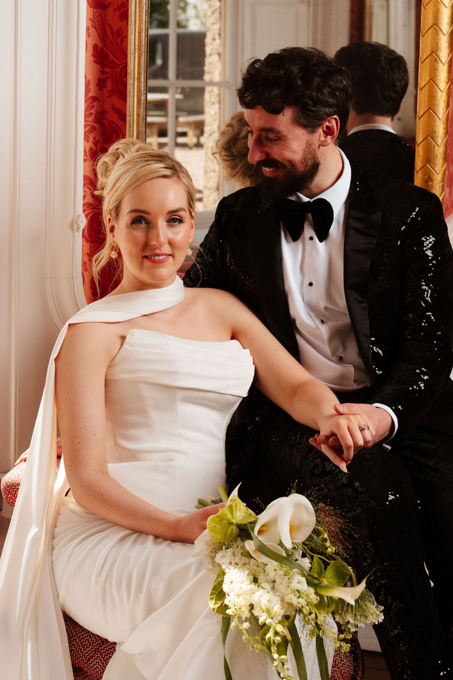 Bride in strapless white gown holding calla lily bouquet seated beside bearded groom in black sequin tuxedo in ornate room.