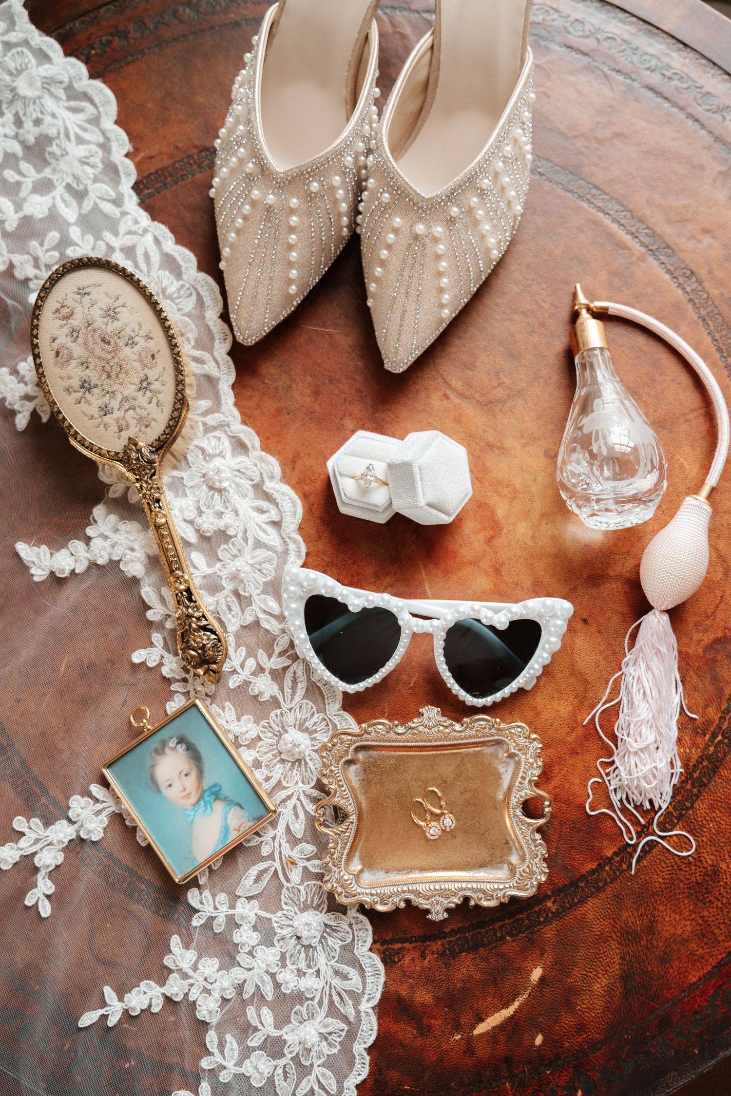 Elegant bridal flat lay with pearl heels, heart sunglasses, perfume, ring box, lace veil, and vintage mirror on brown surface.
