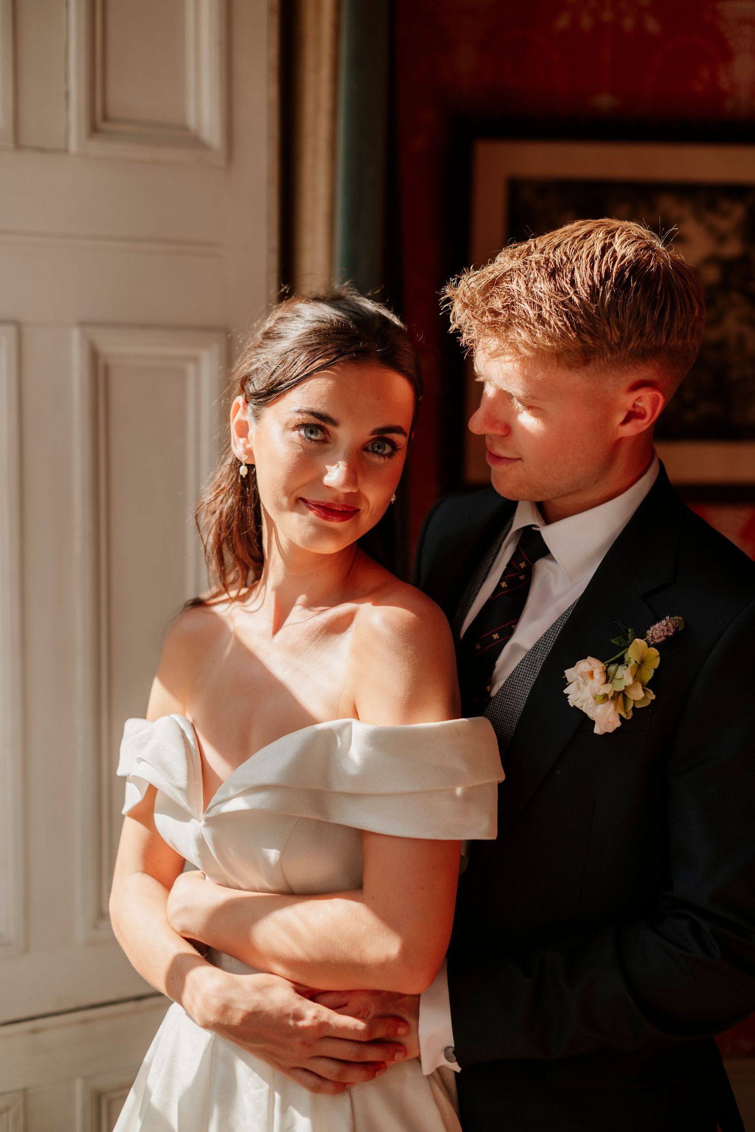 A bride in an off-shoulder white gown embraces her groom in a black suit with floral boutonniere indoors.