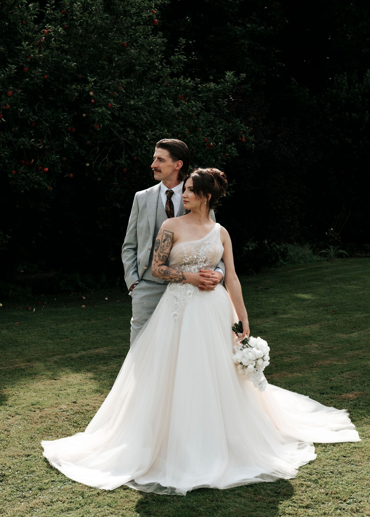 Bride in lace ball gown and tattooed groom in grey suit pose together in lush green garden setting.