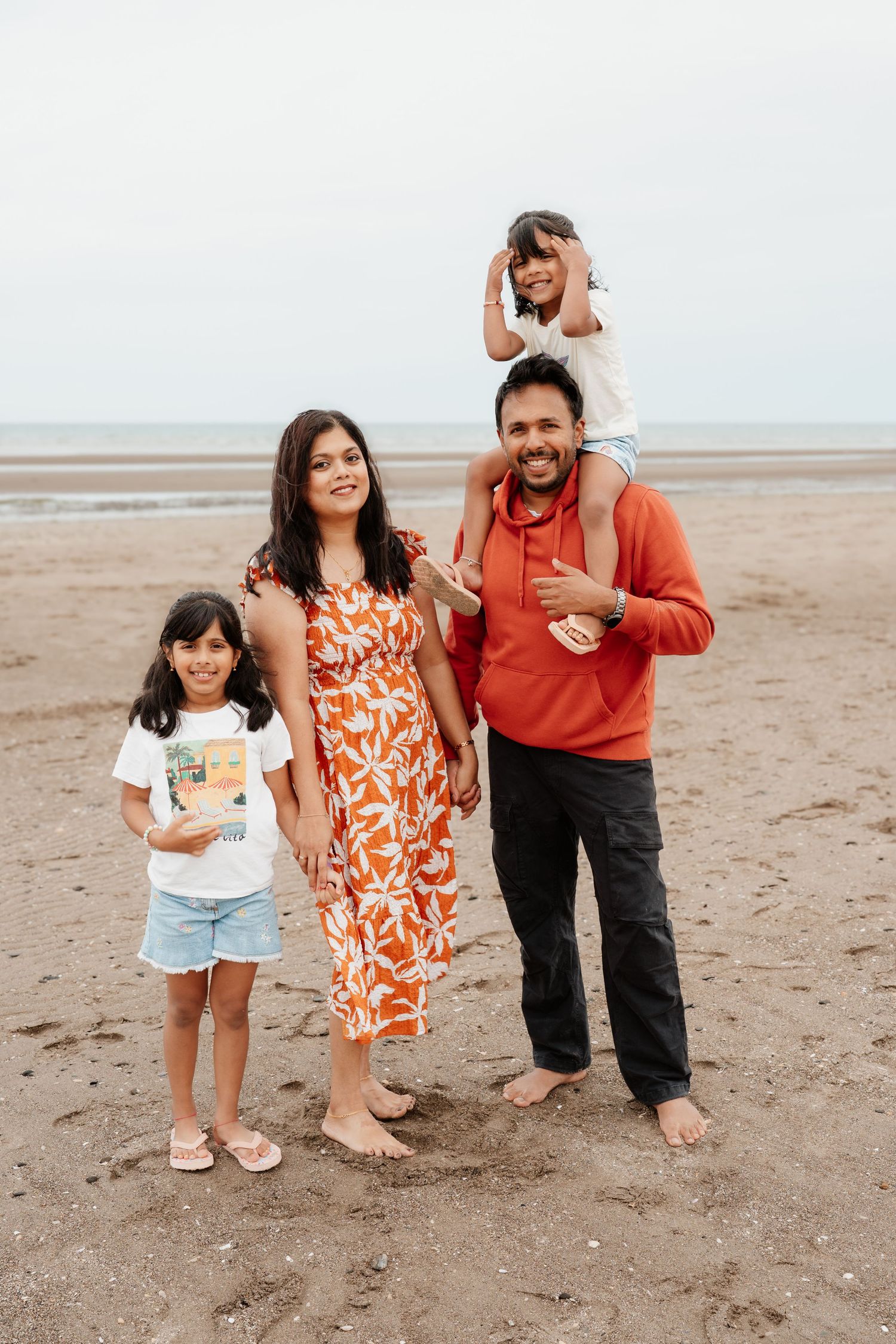 Happy South Asian family of four posing on a sandy beach, parents with two young daughters, wearing casual colorful outfits.