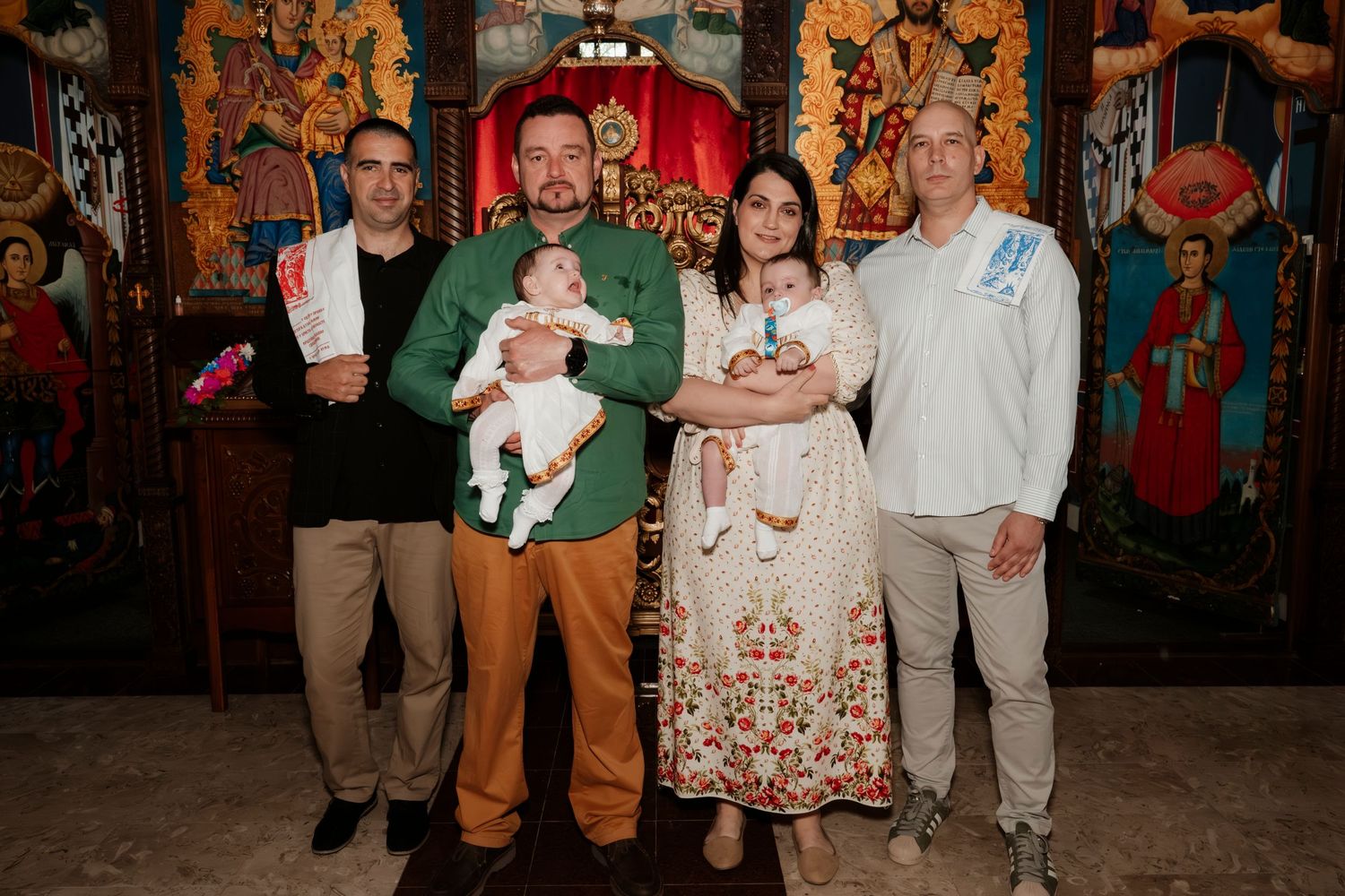 Family posing with babies in front of ornate Orthodox church iconostasis during a baptism ceremony.