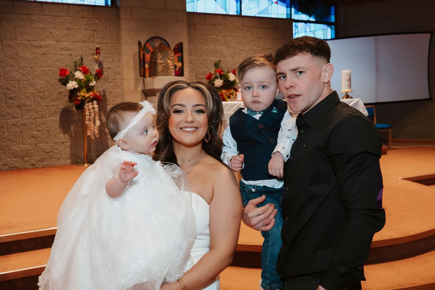 Family posing in church during christening ceremony, mother in white dress holding baby girl in white gown with toddler son.