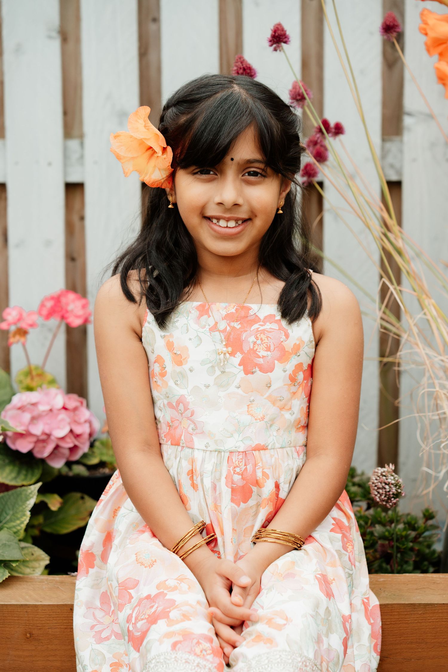 Young girl in floral dress with orange flower in hair smiling outdoors near garden fence.