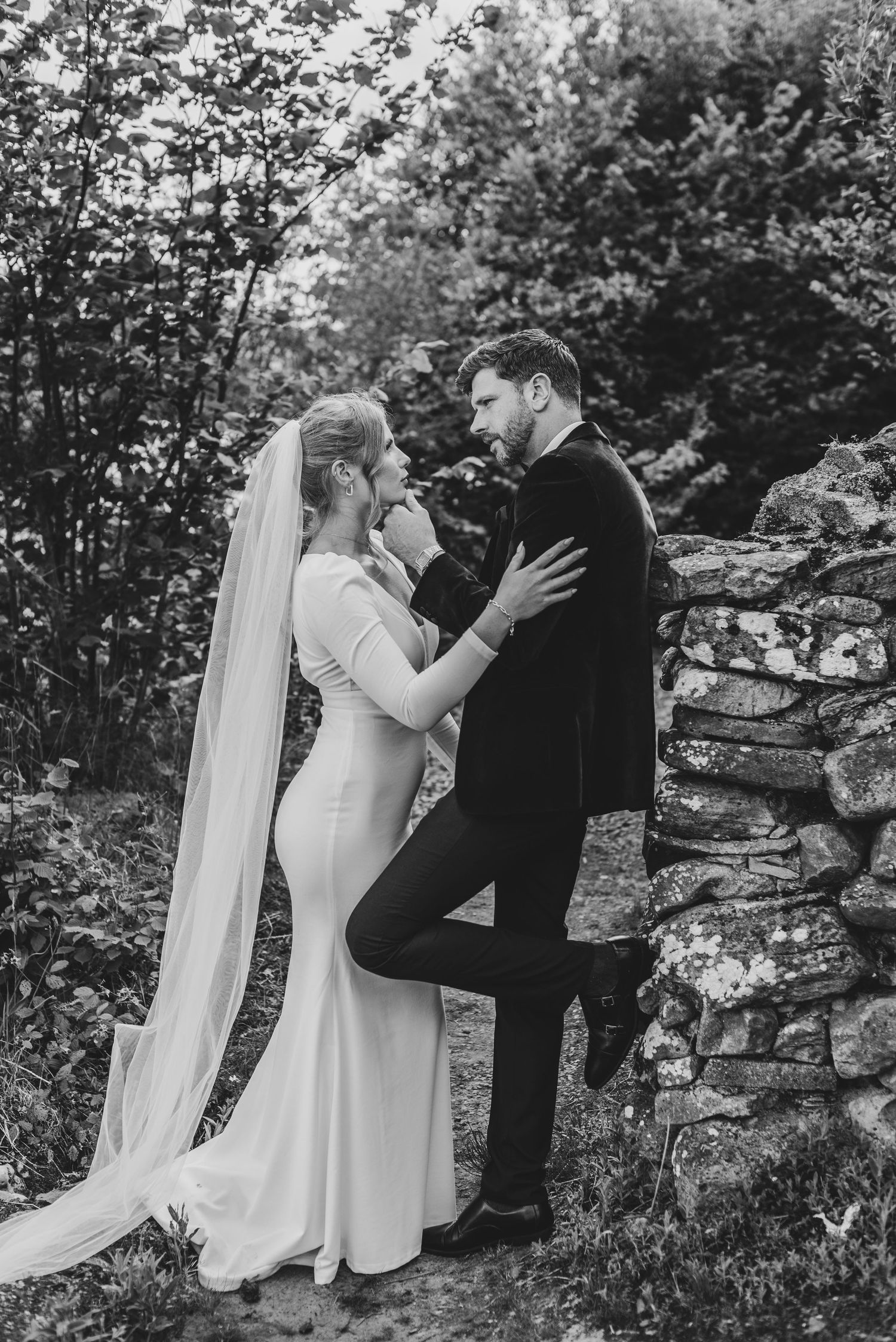 Black and white wedding portrait of bride and groom sharing intimate moment by rustic stone wall.