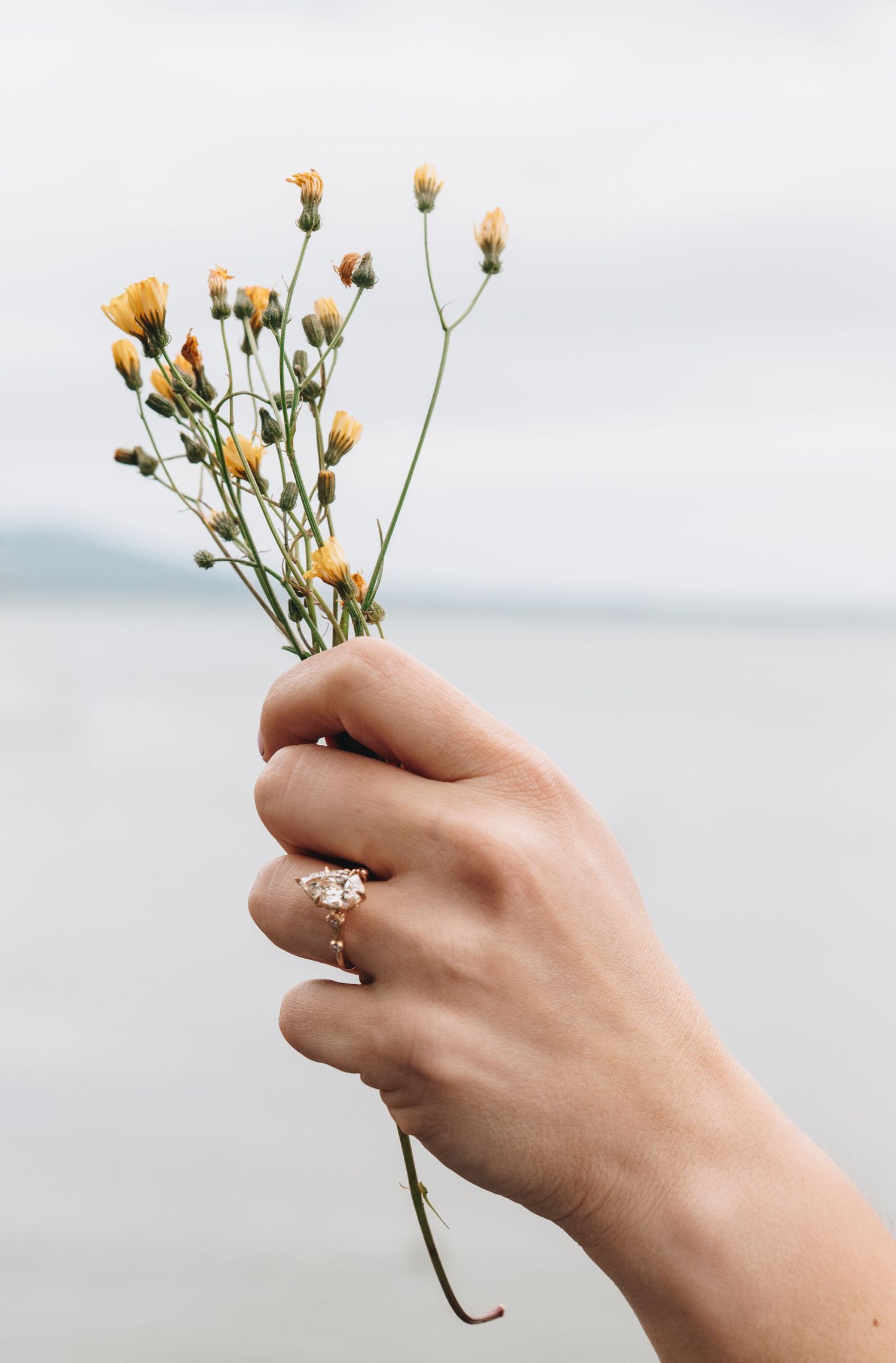 Hand holding small yellow wildflowers with a gold oval engagement ring against a soft ocean backdrop.
