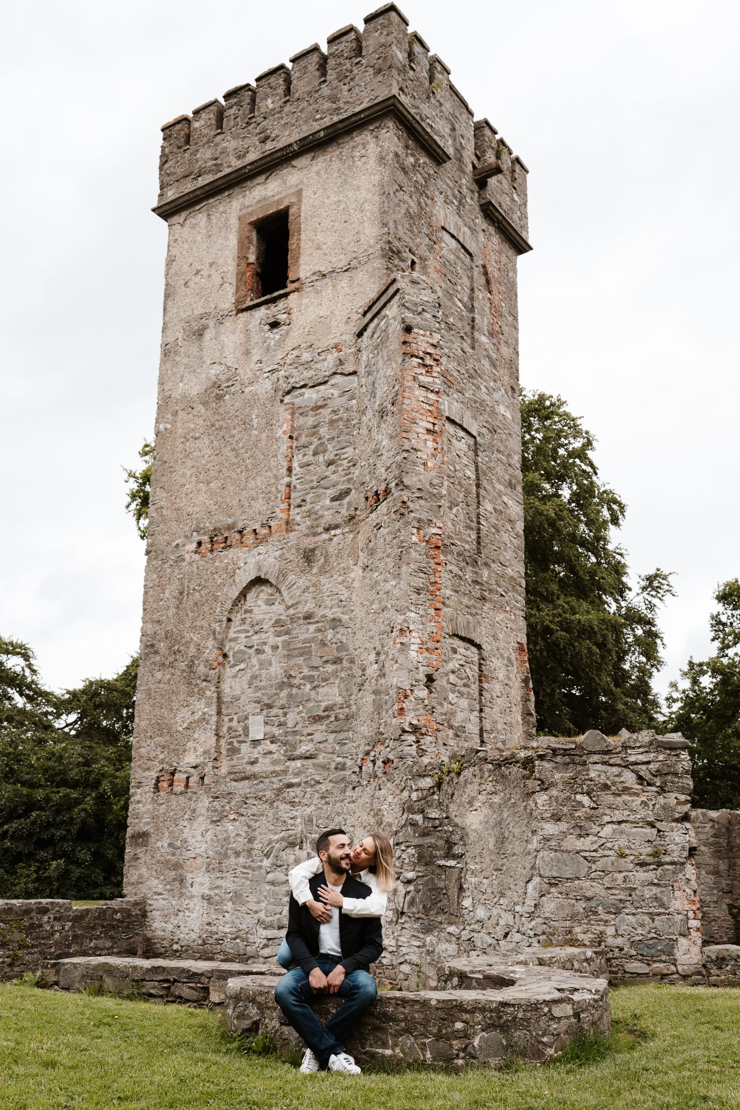 Couple sitting romantically before an ancient stone castle tower ruin on green grass under overcast skies.