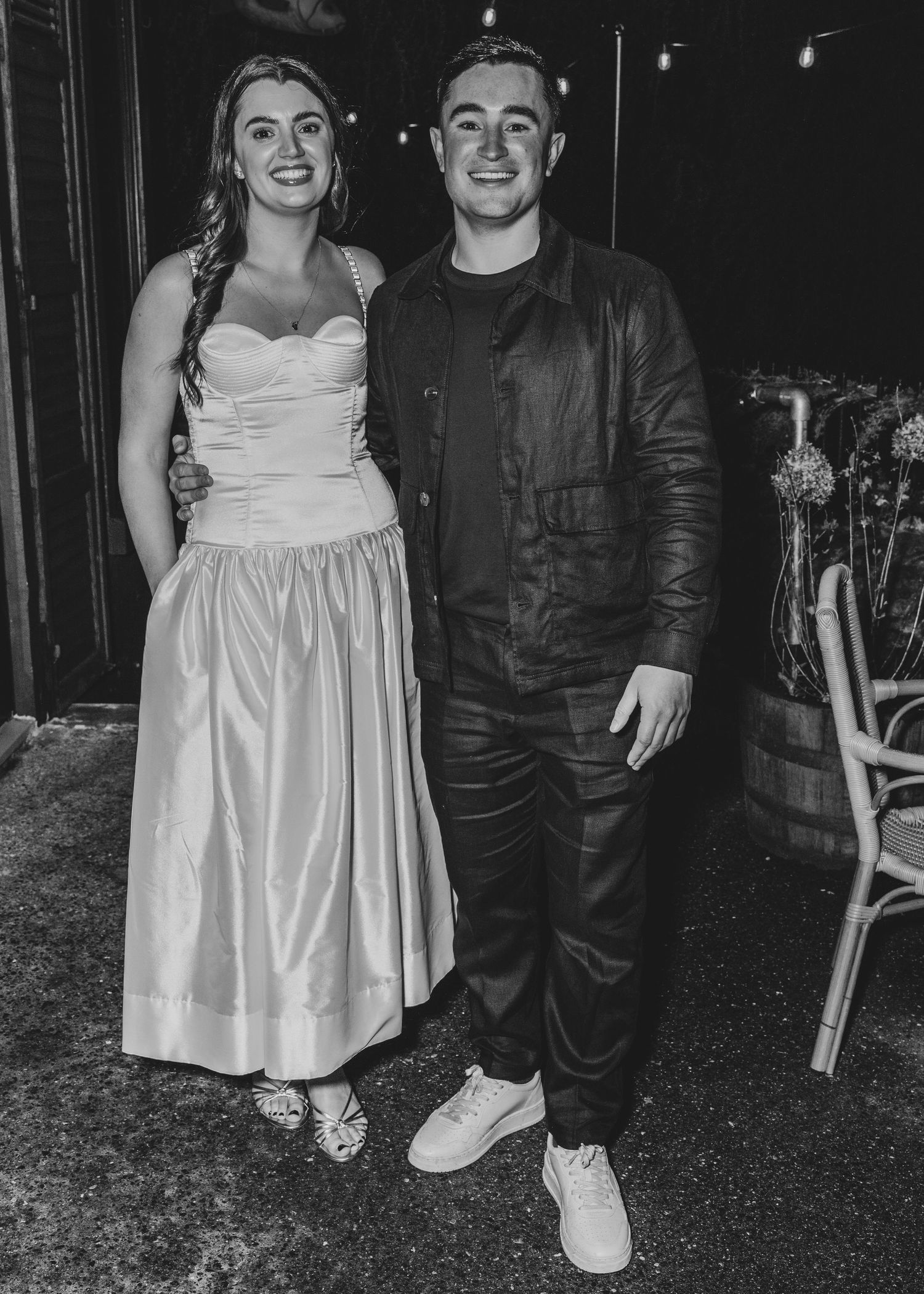 Black and white photo of a smiling couple posing together, woman in elegant gown and man in leather jacket.