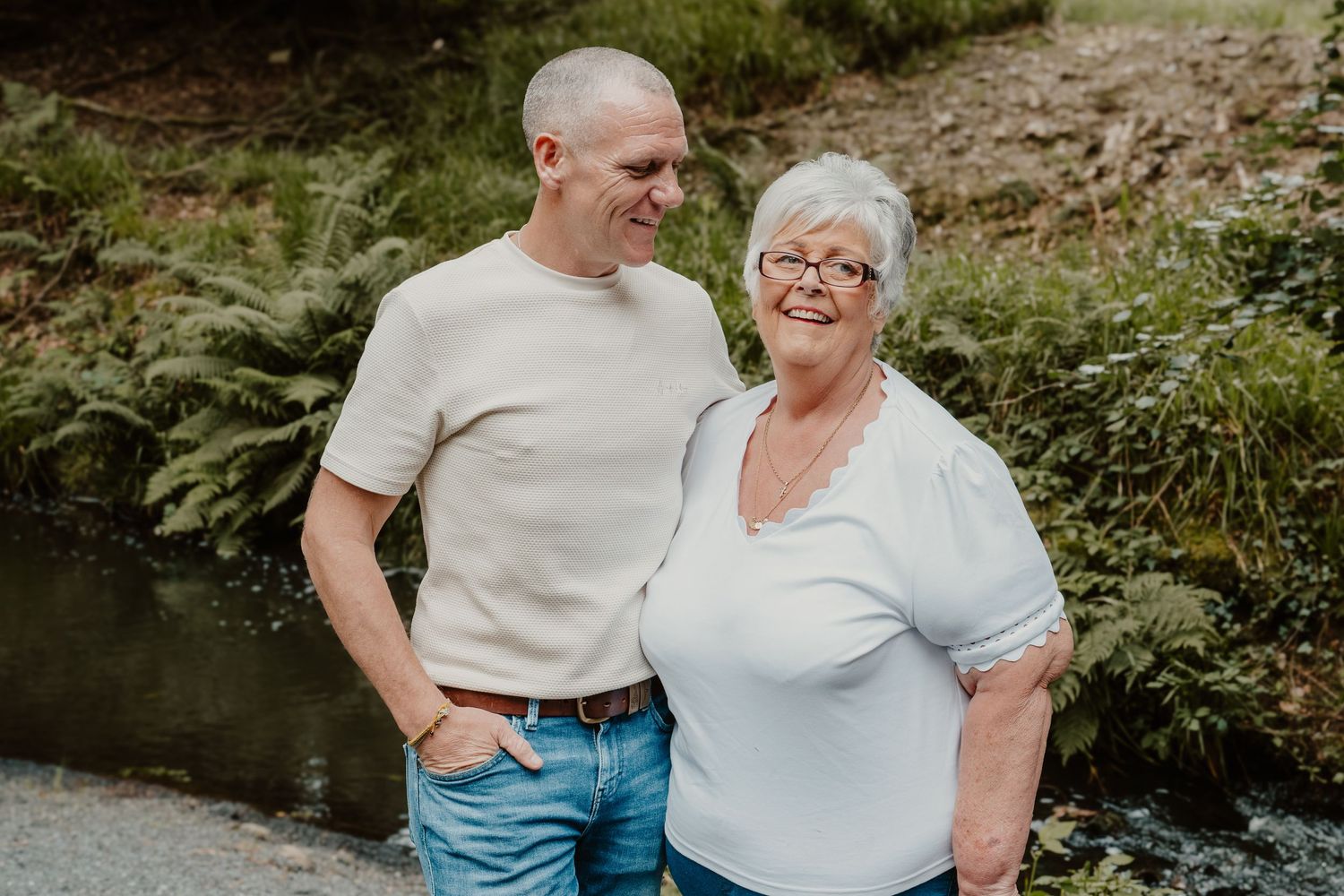A man and older woman smiling together outdoors near a stream surrounded by lush green ferns.