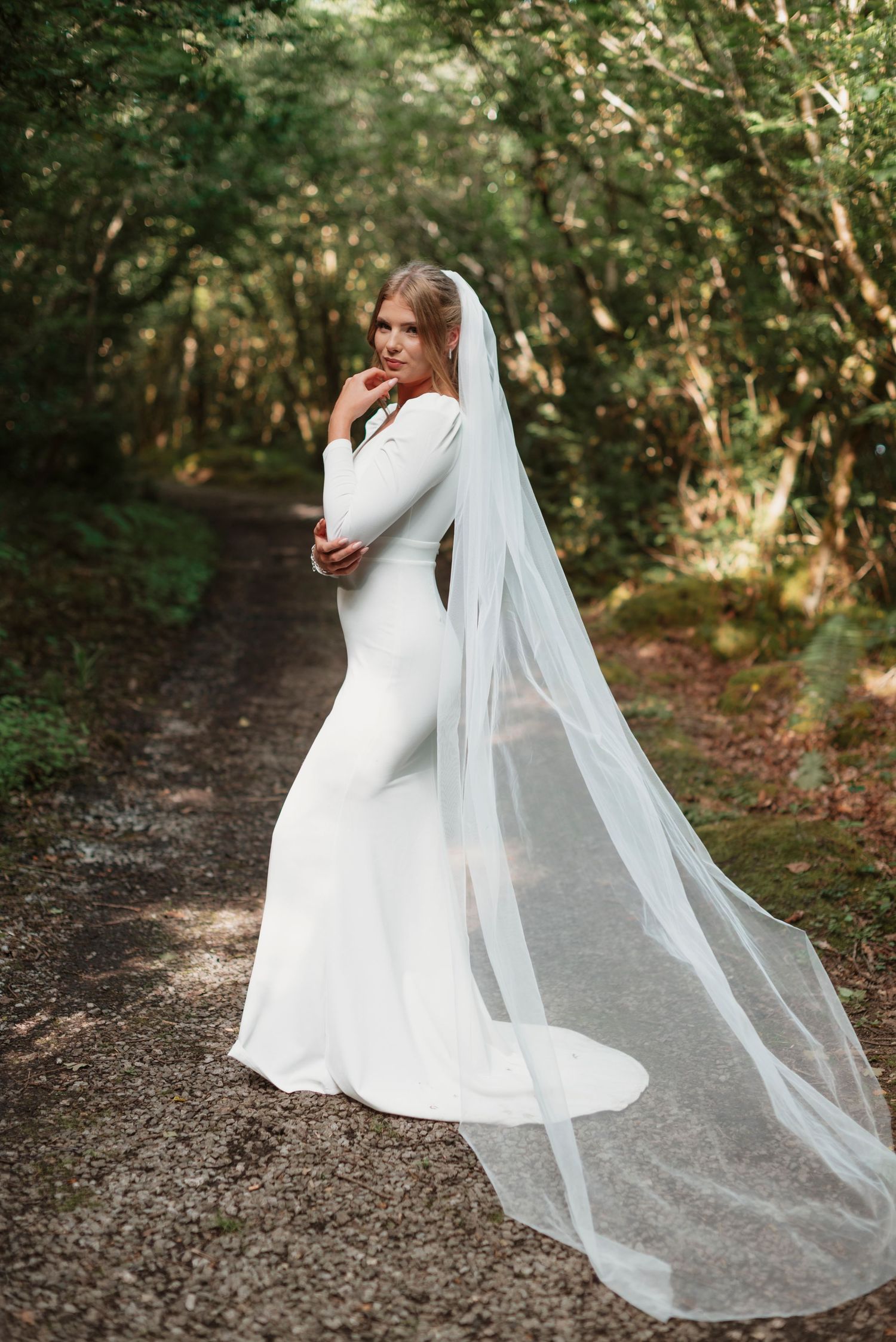 Bride in fitted long-sleeve white wedding gown with dramatic cathedral veil poses on forest path.
