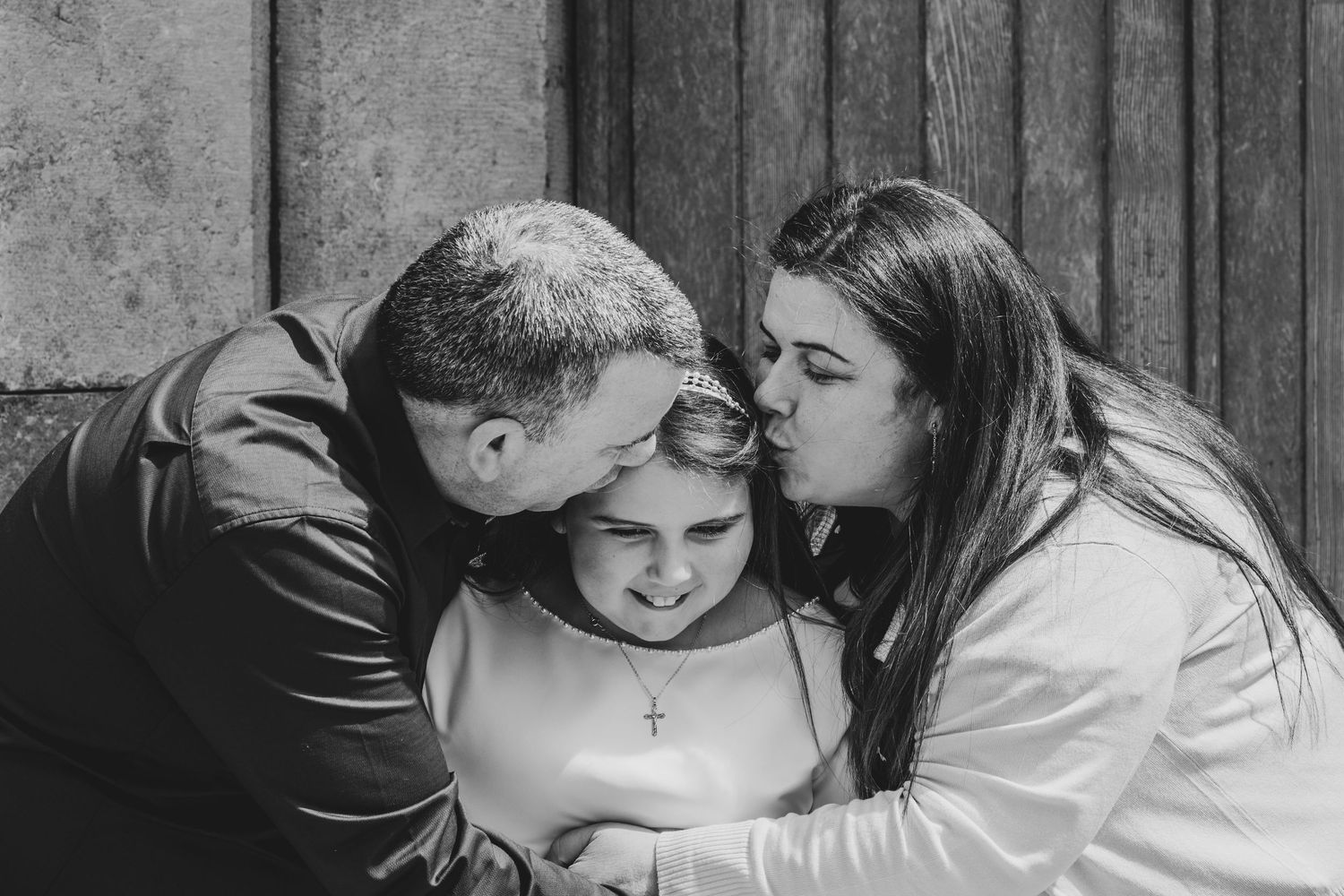 Black and white photo of parents kissing their young daughter on the cheeks, child wearing cross necklace.