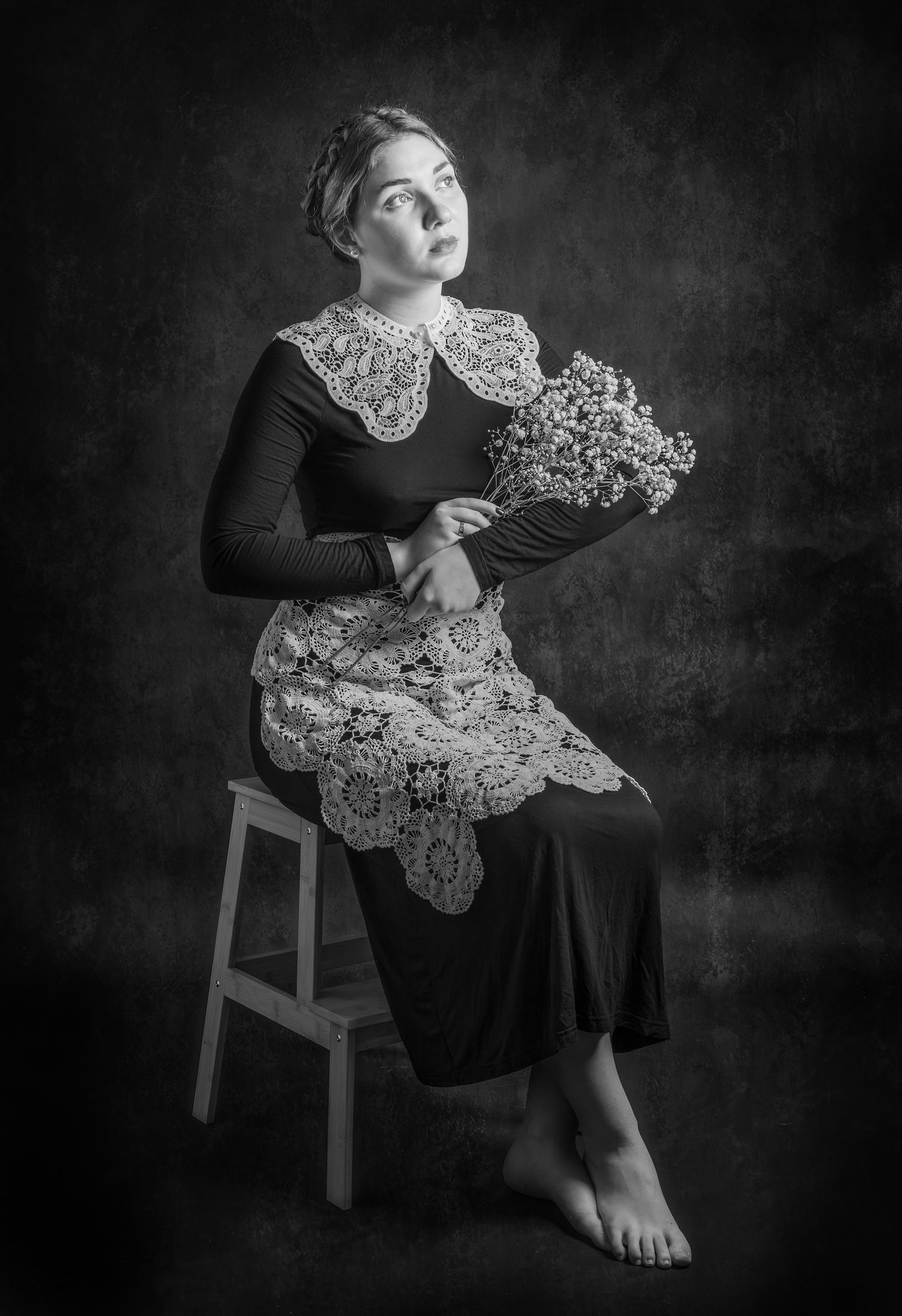 Black and white portrait of a young woman seated on a stool, wearing lace collar and apron, holding dried flowers.