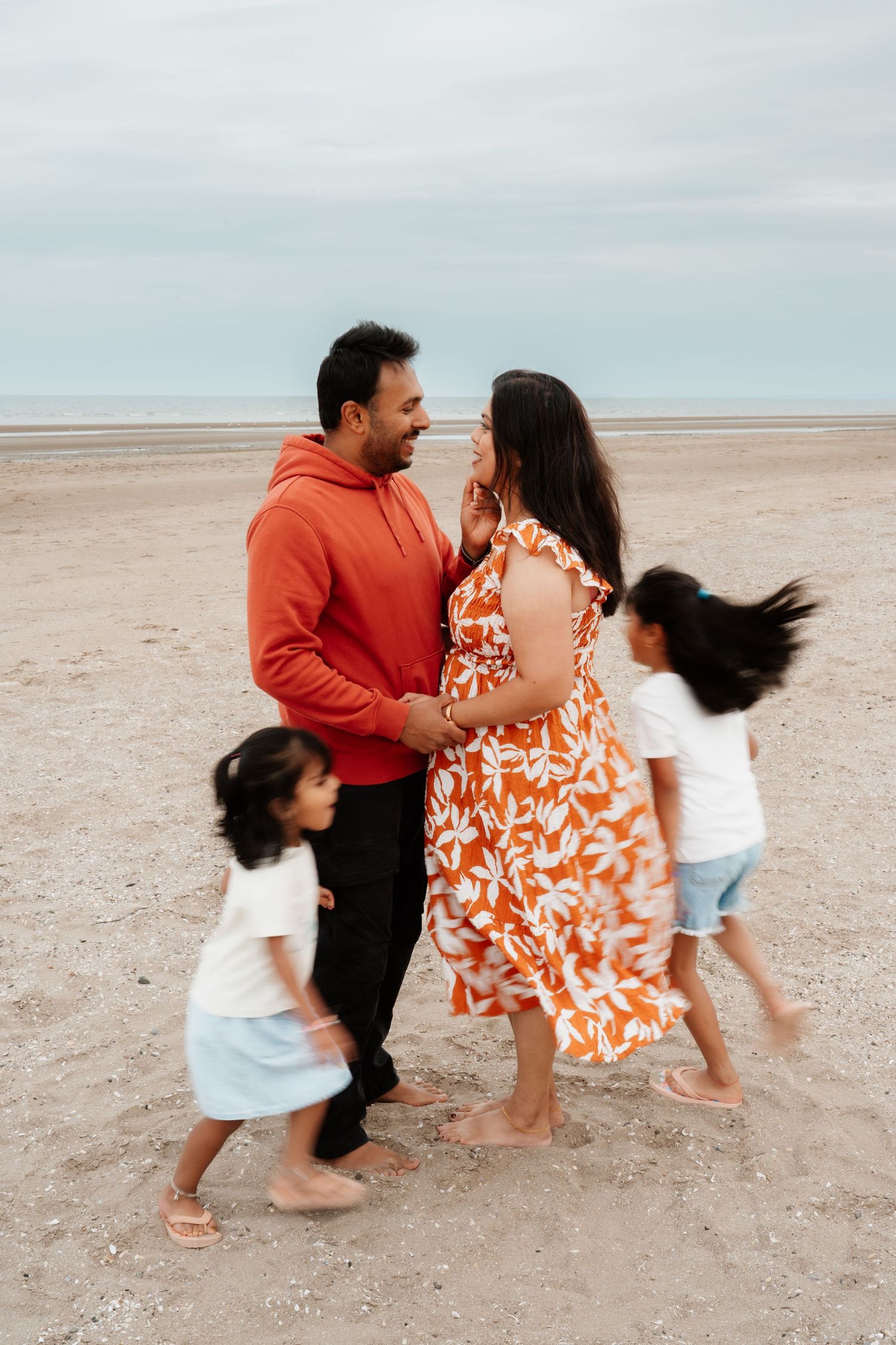 A family of four enjoys a beach day, with parents sharing a tender moment while two young children play around them.