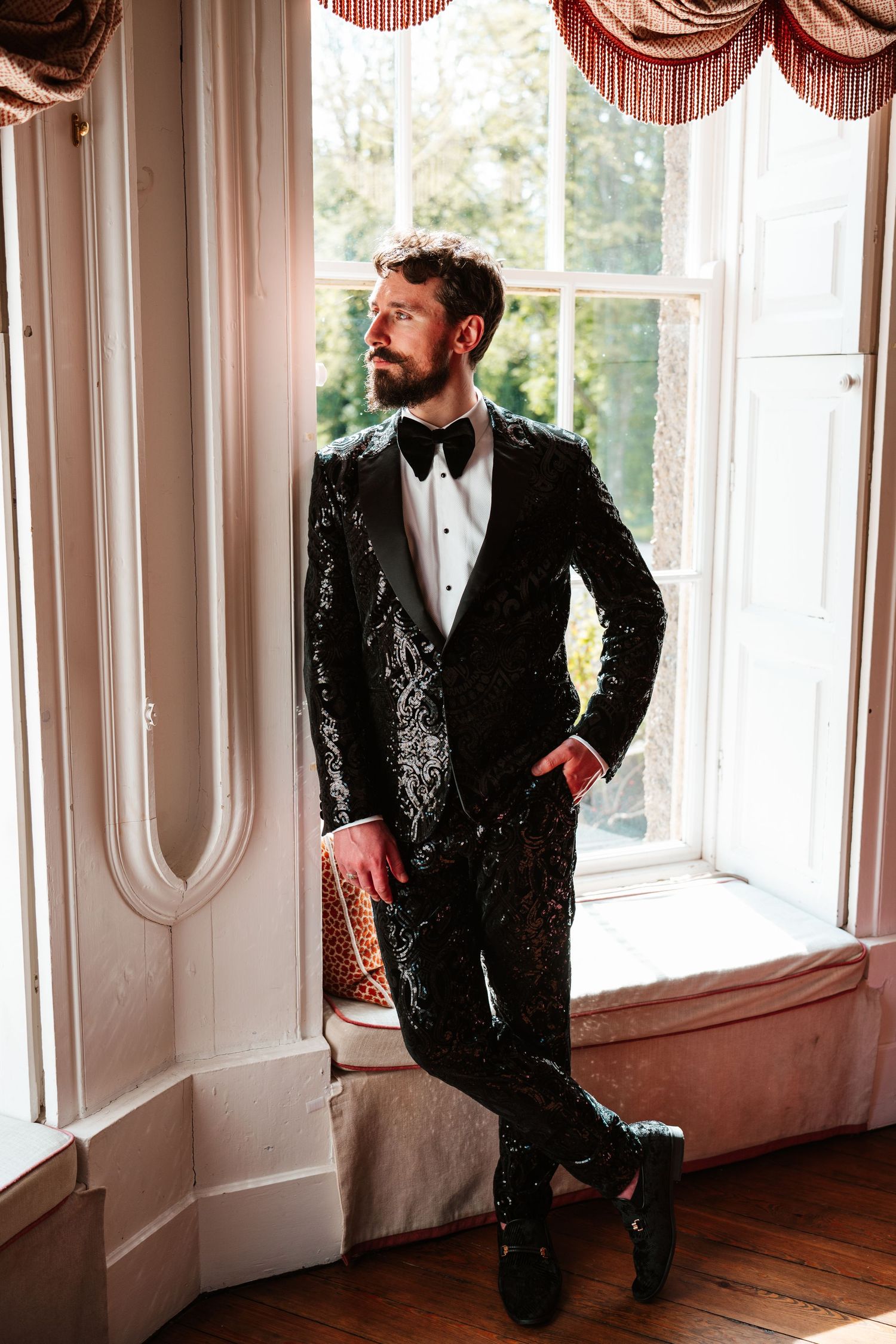 Groom in black sequin tuxedo with bow tie leans against window in elegant interior setting.