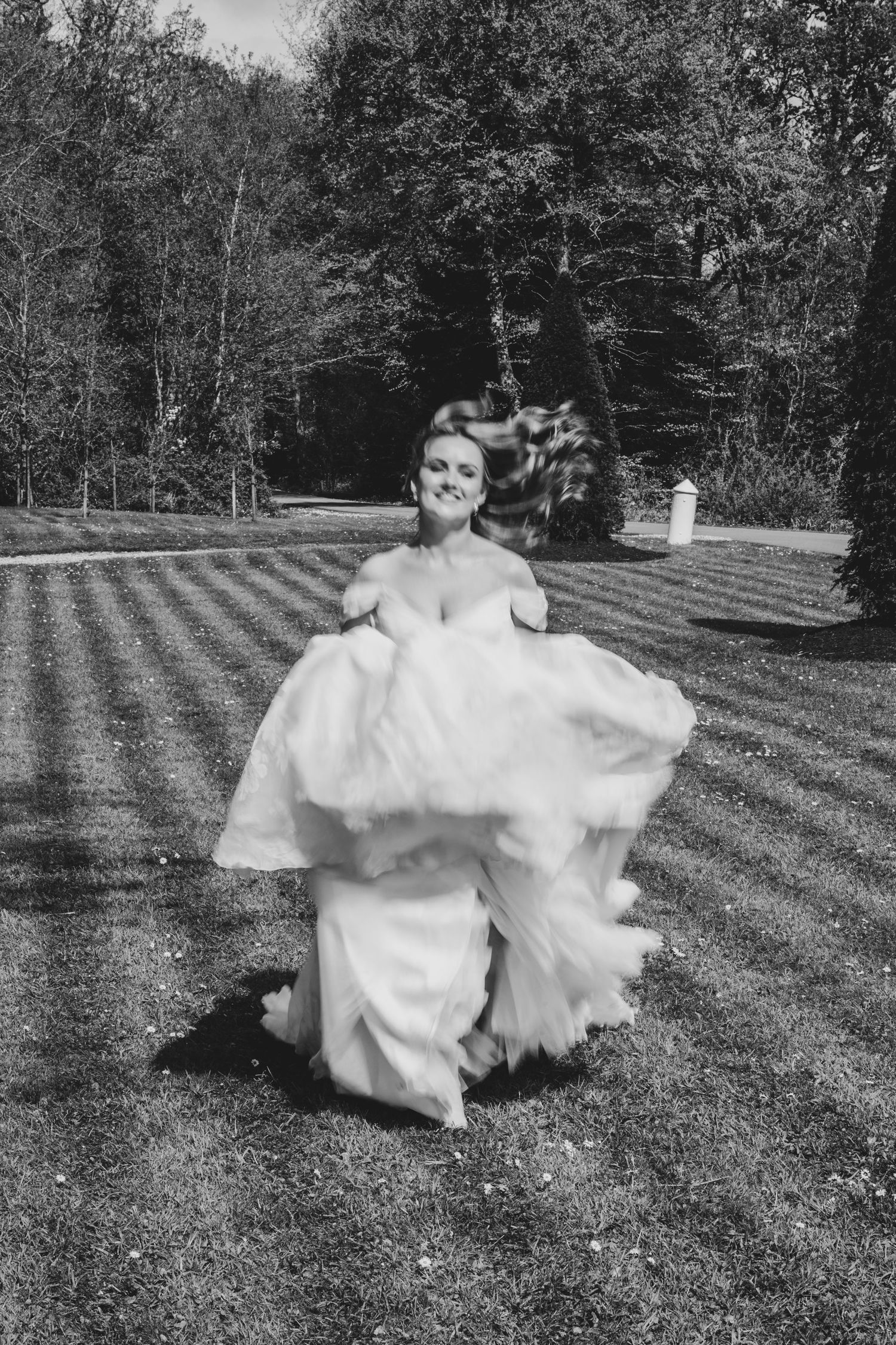 Bride in flowing white gown running joyfully across striped lawn, black and white wedding photo outdoors.